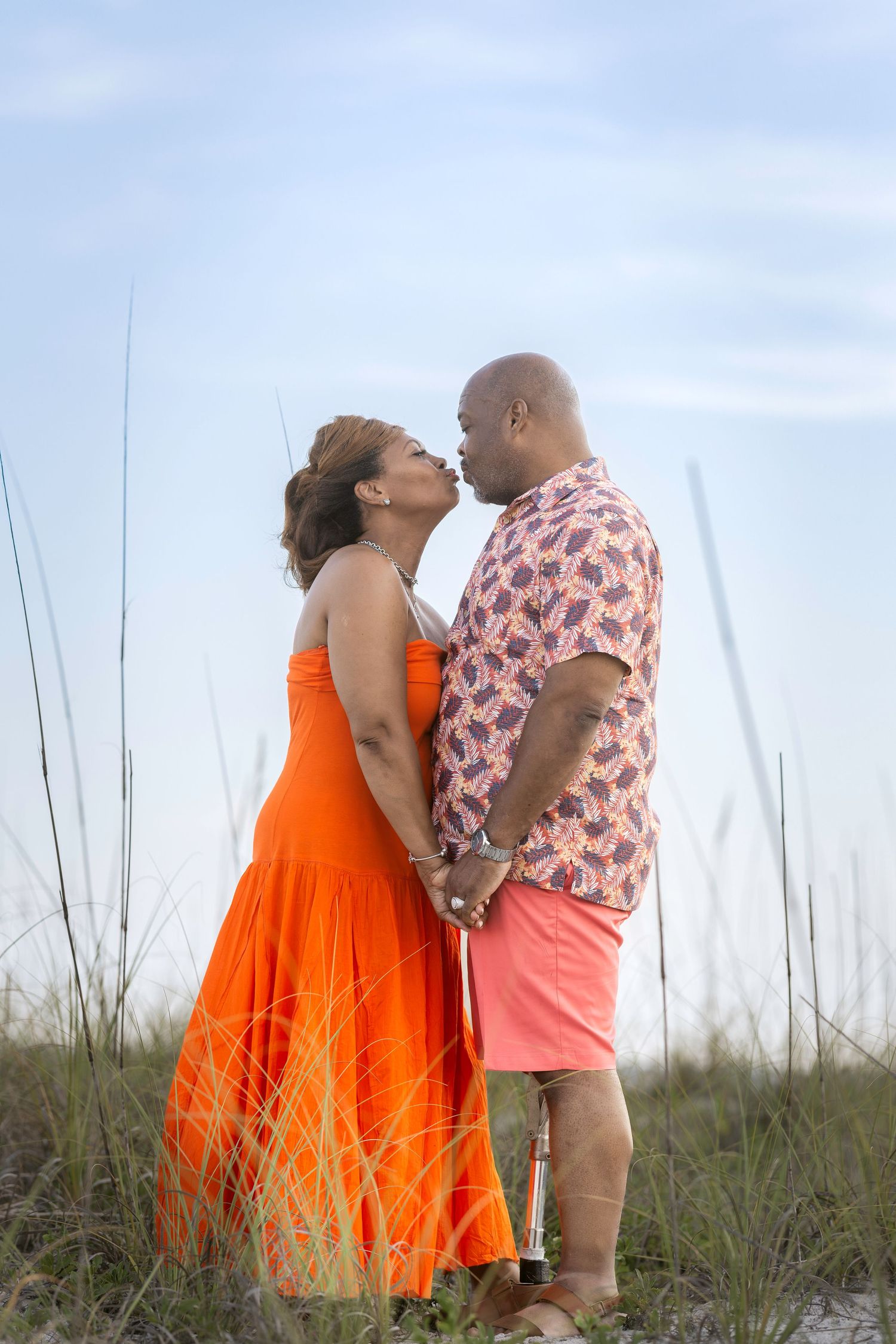 A couple shares a romantic kiss on a beach, woman in orange dress, man in pink floral shirt holding a bottle.