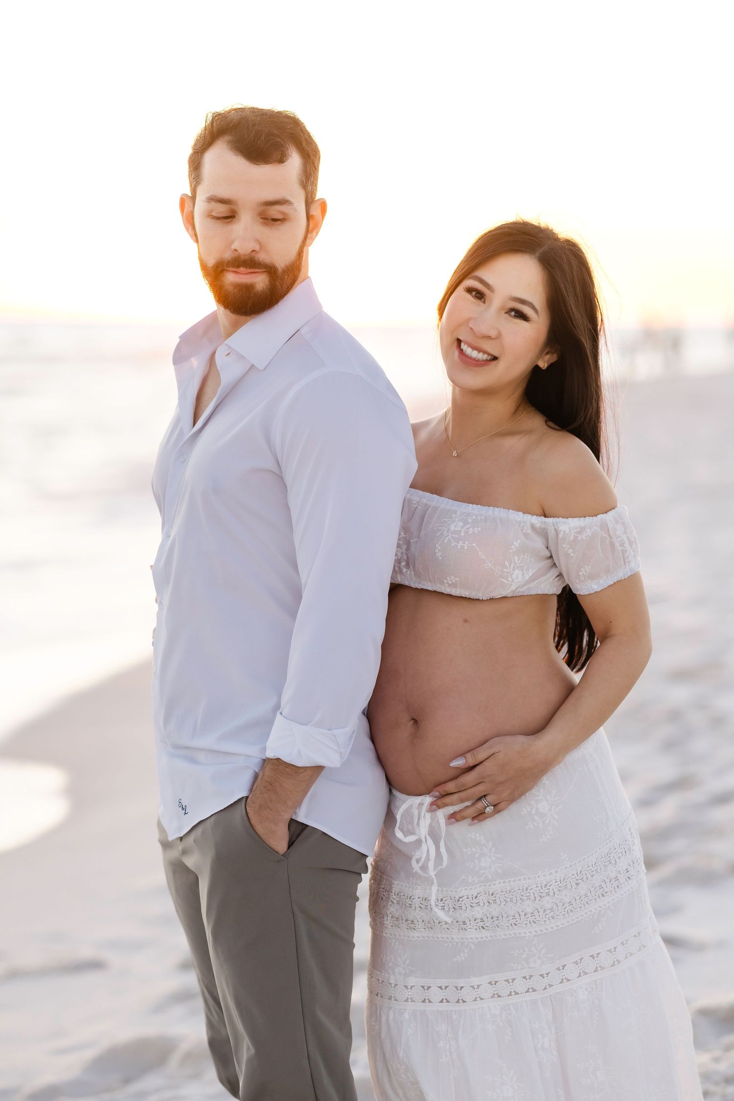 A couple shares intimate moments on a bright sandy beach during a romantic maternity photoshoot.