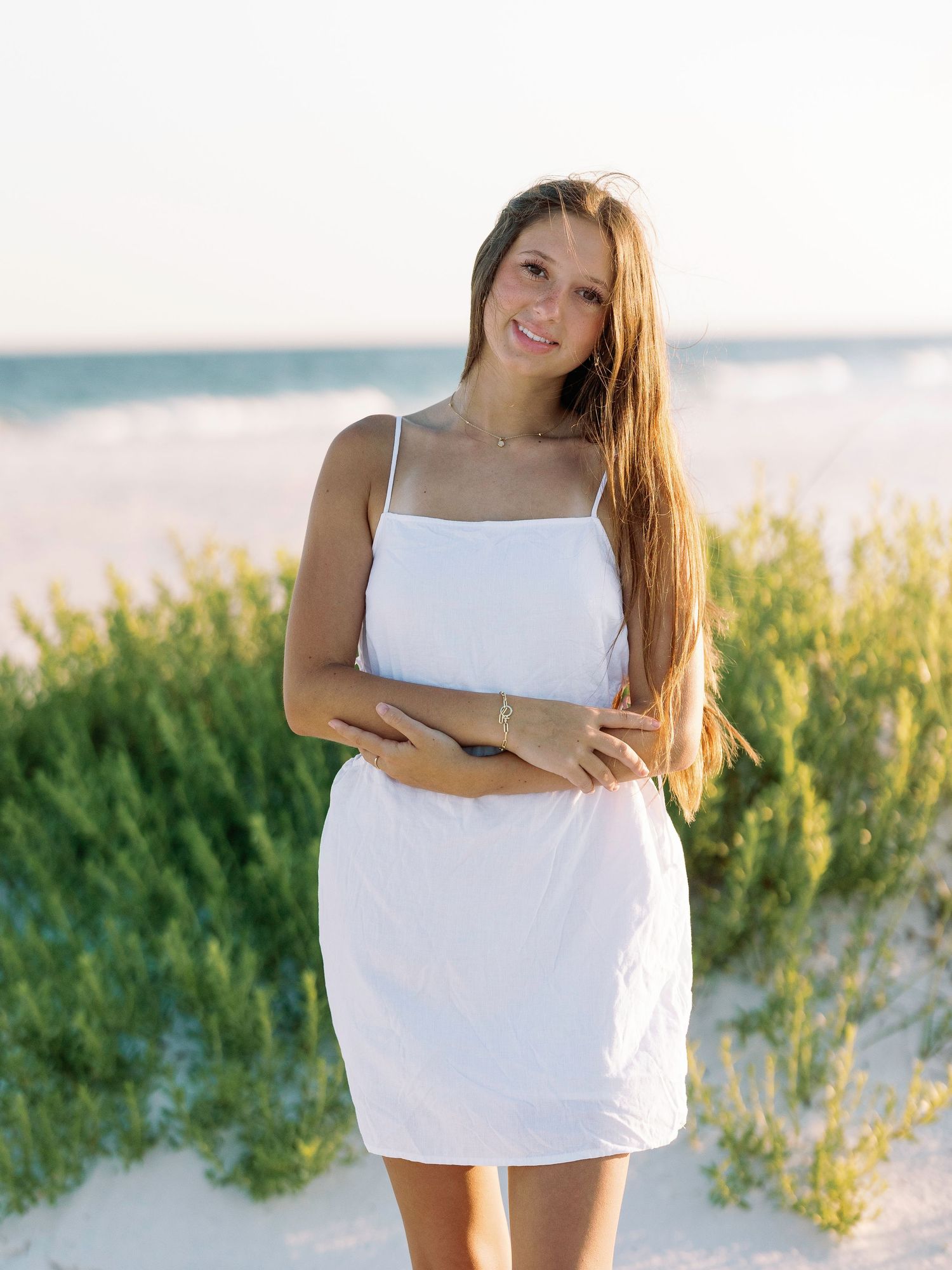 Woman in white sundress poses on sandy beach with coastal vegetation in background.