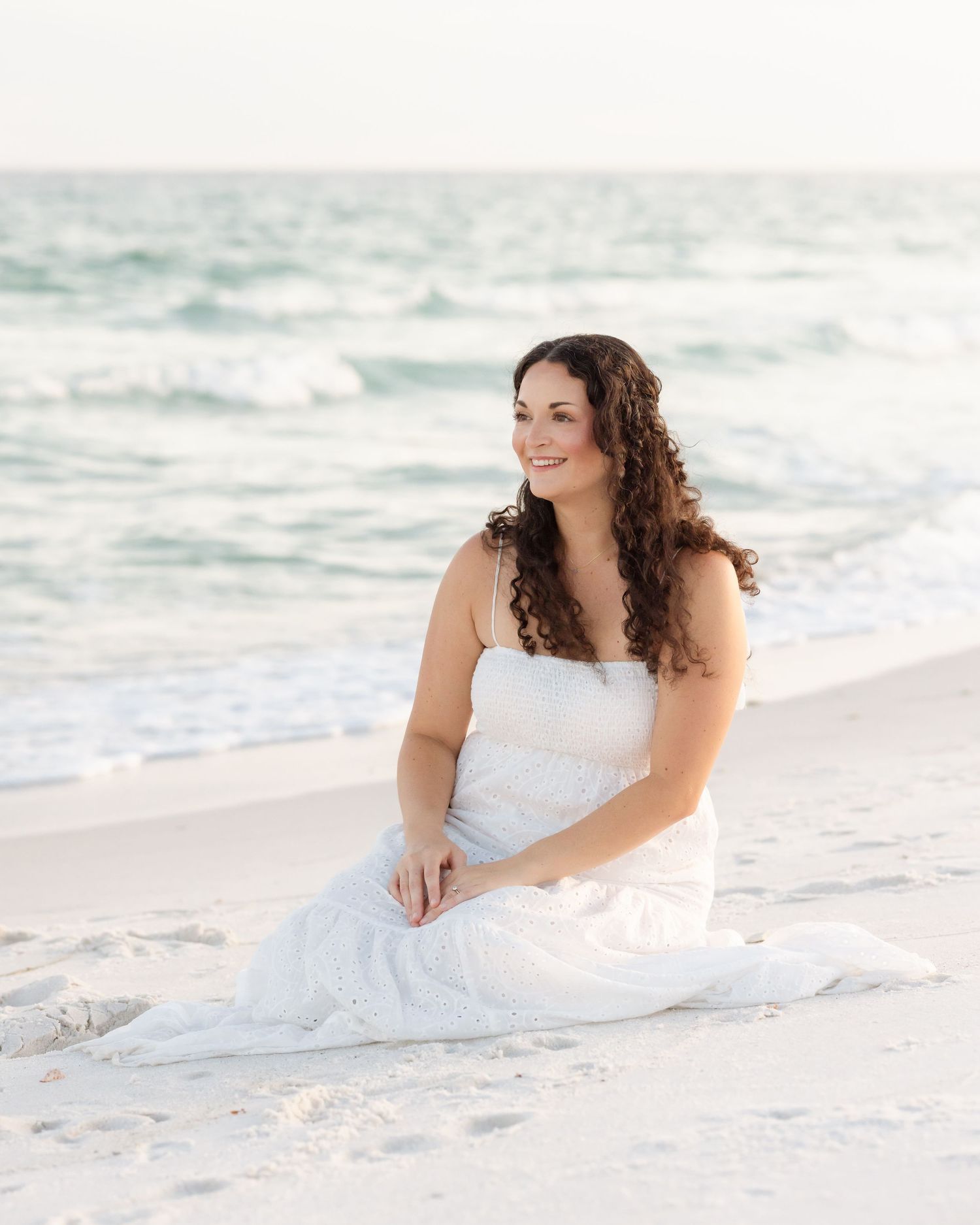 Woman in white dress sitting gracefully on sandy beach with turquoise ocean waves in background.