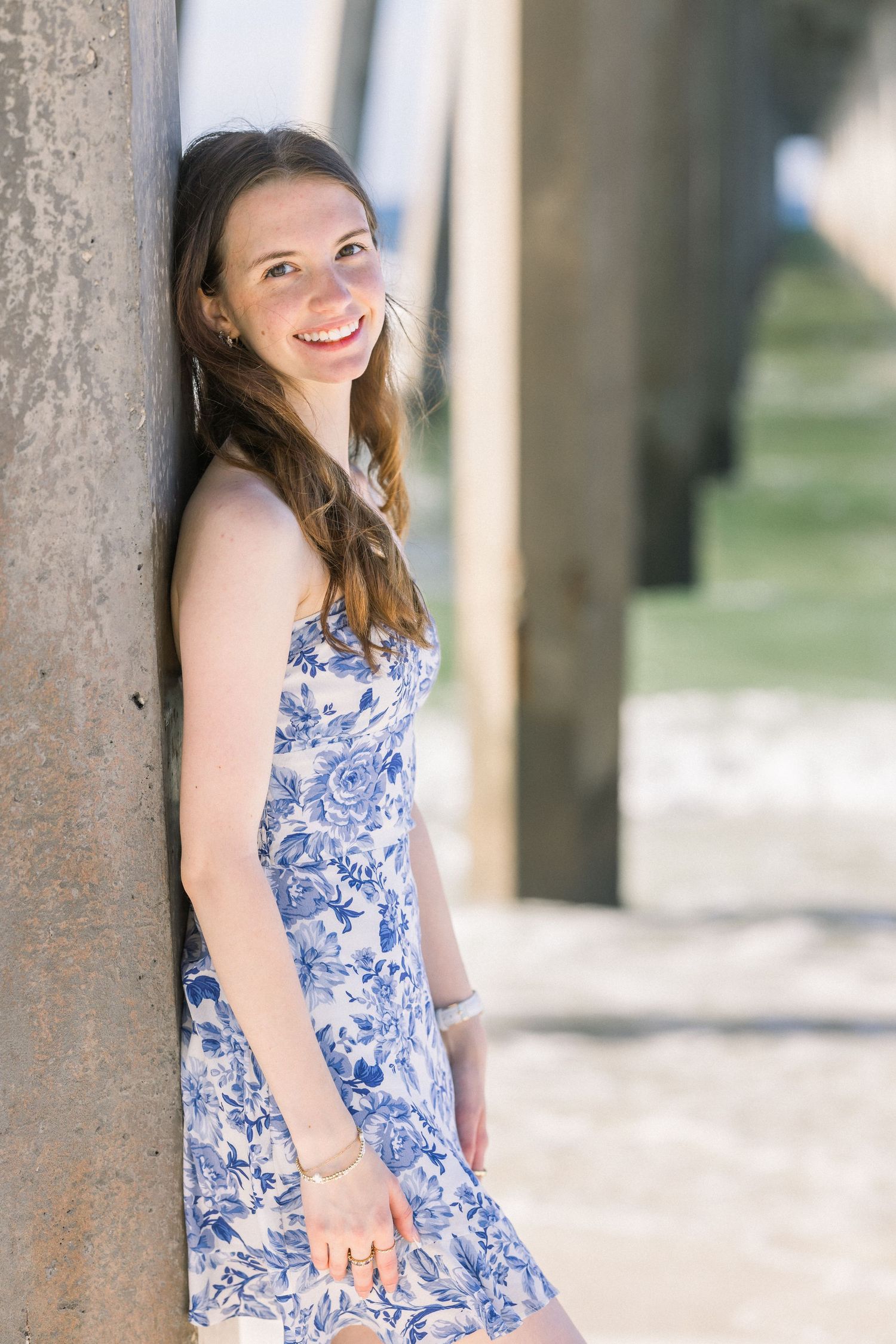 Young woman in blue floral sundress smiling and leaning against concrete pillar outdoors on sunny day.