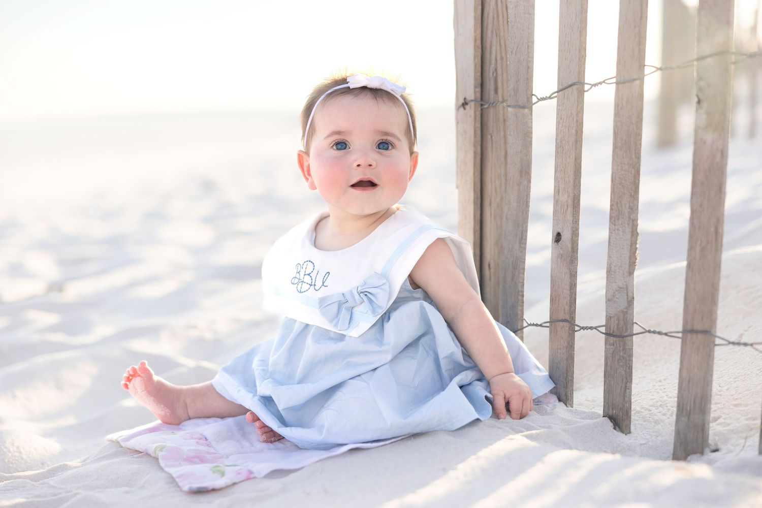 Baby in white dress sitting on sandy beach with wooden fence during sunset creates dreamy coastal scene.