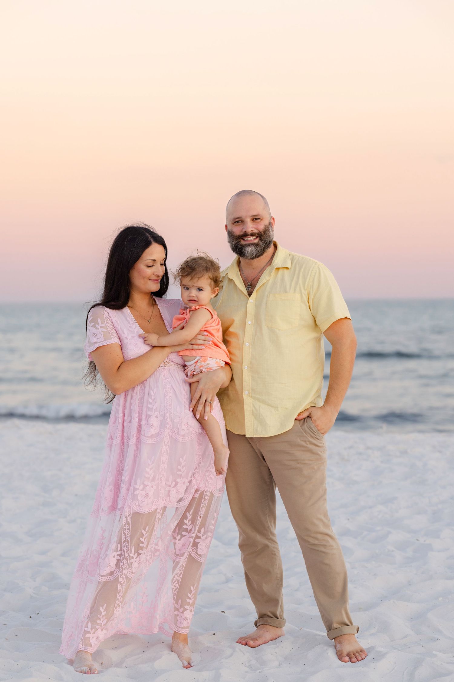 A family enjoys a sunset beach photoshoot wearing pastel colors while standing in the white sand.