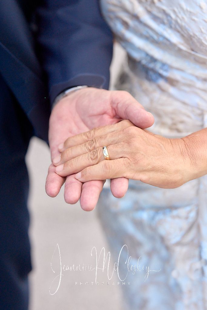 Close-up of hands showing wedding rings with bride in white dress and groom in navy suit holding hands together.