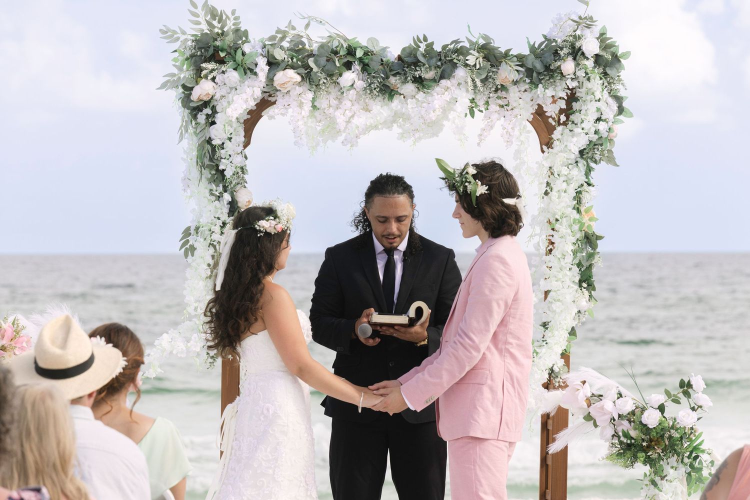 Two brides exchange vows at a beach wedding ceremony under a floral arch, officiated by a man in a black suit.
