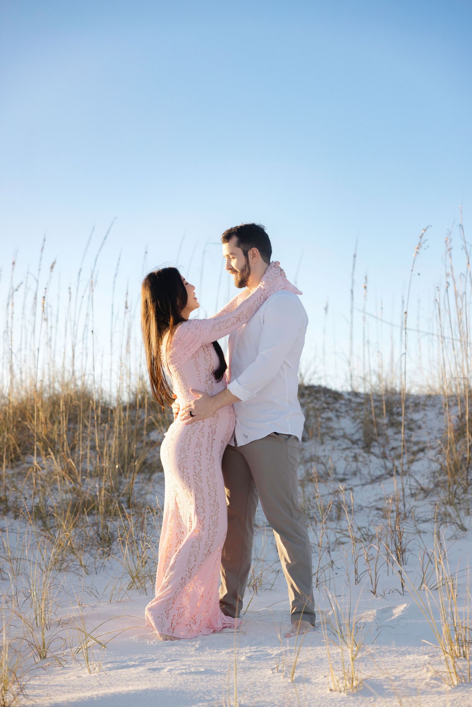 Couple embracing on white sand dunes with sea oats, woman in pink gown, man in white shirt, blue sky background.