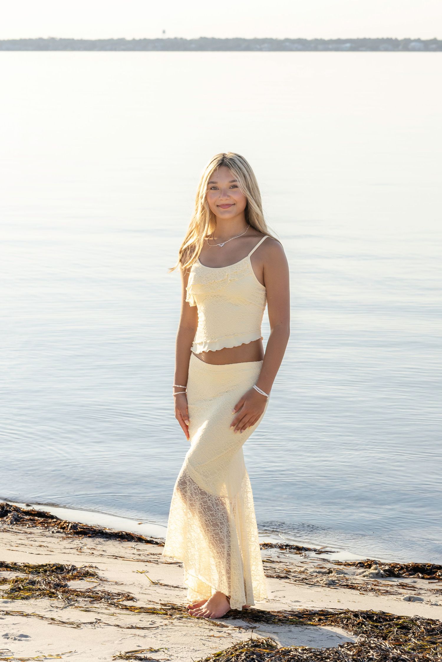 A beachside photoshoot featuring someone in a cream-colored outfit posing against the ocean and sand backdrop.