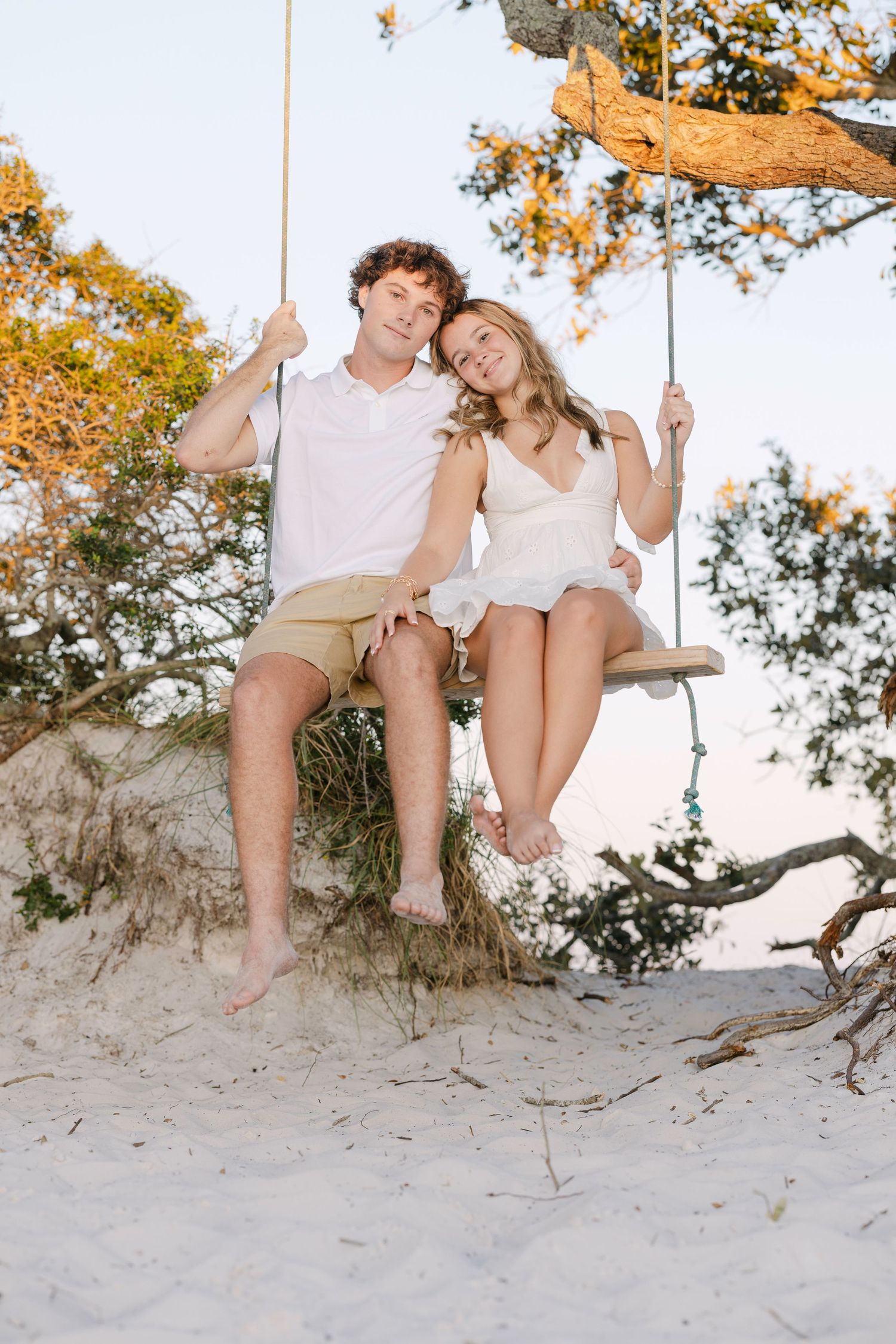 Two people in summer outfits share a romantic moment on a wooden swing at a sunny beach with golden fall trees.