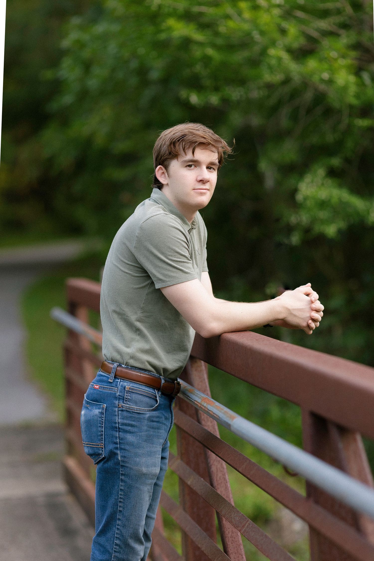 Person in gray t-shirt and jeans leans against wooden railing on outdoor path surrounded by greenery.