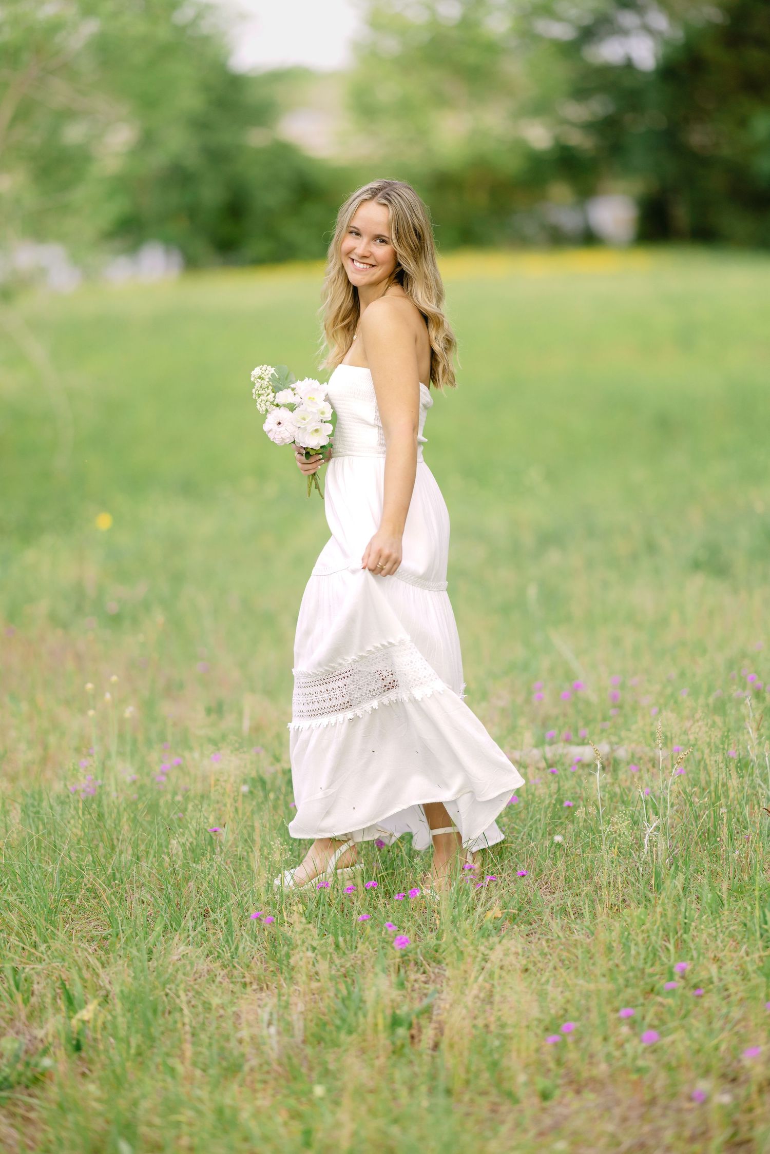 A woman in a flowing white dress twirls gracefully in a green meadow while holding a delicate bouquet of flowers.