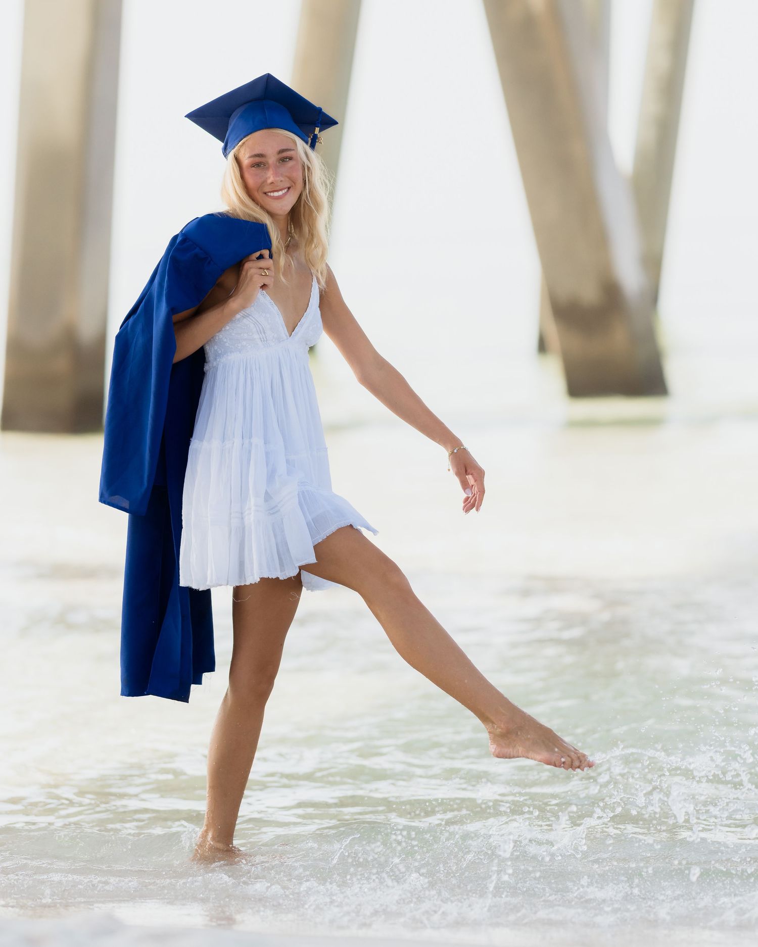 Smiling blonde graduate in white dress and blue cap and gown posing barefoot on a sunny beach near a pier.