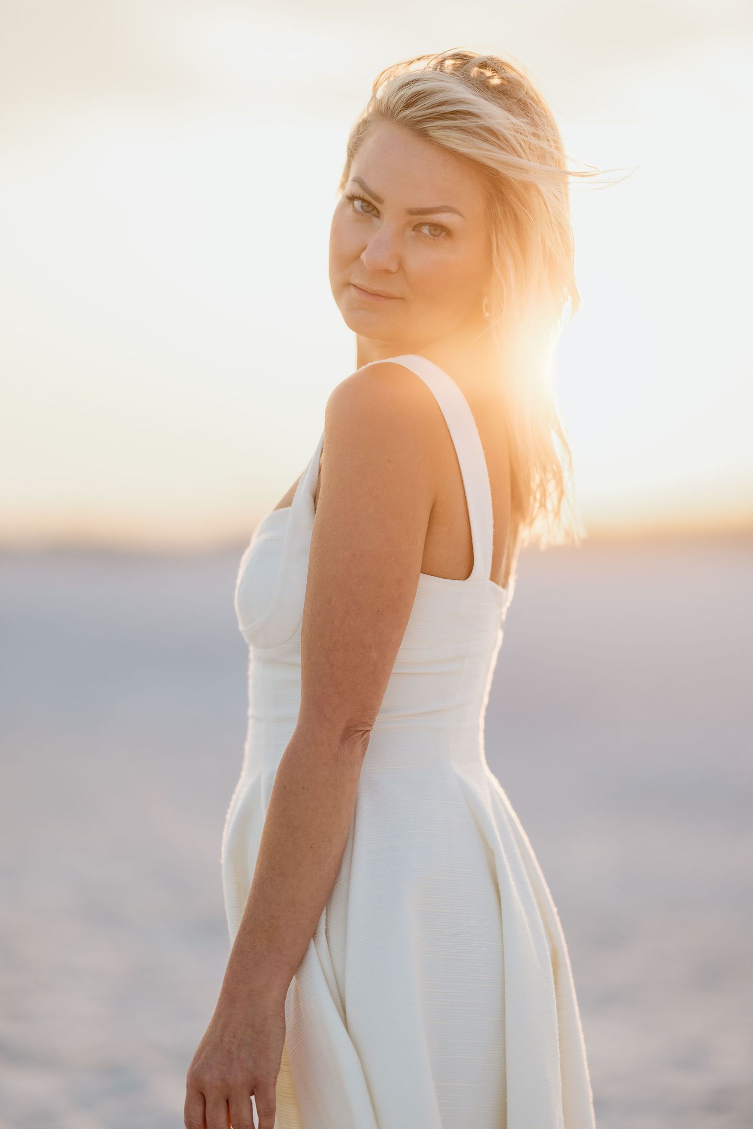 Woman in white sundress standing on sandy beach at golden sunset with wind-blown blonde hair.