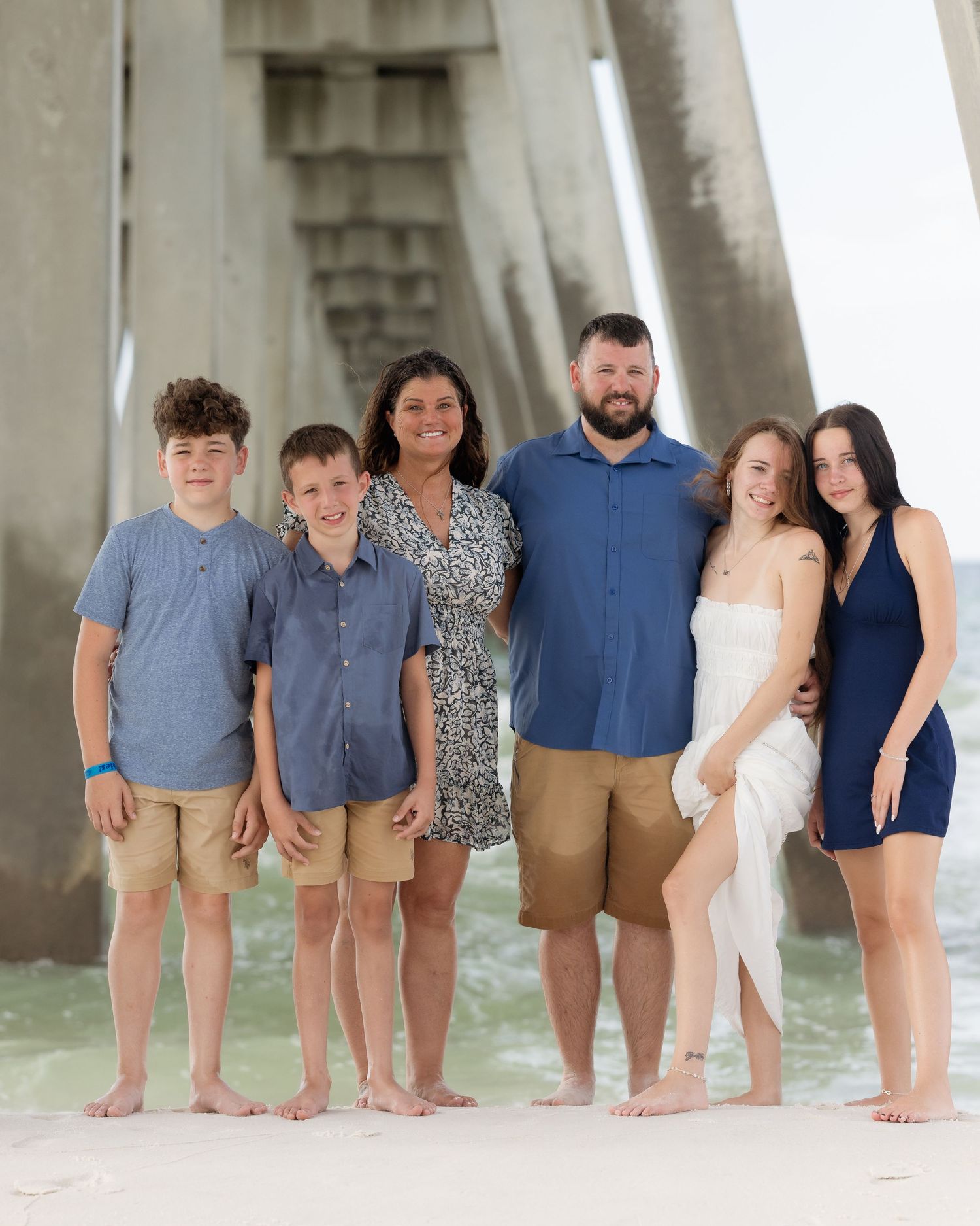 Happy family of six posing barefoot under beach pier, wearing coordinated blue and tan outfits.