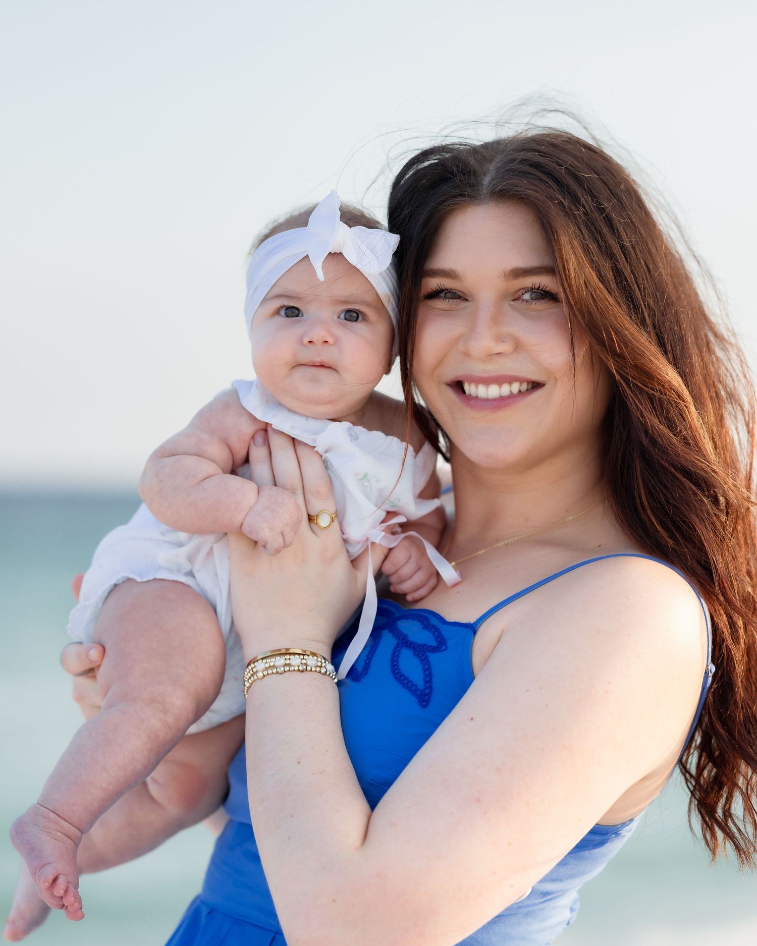 Young woman in blue dress holds baby girl with white bow headband on a sunny beach.