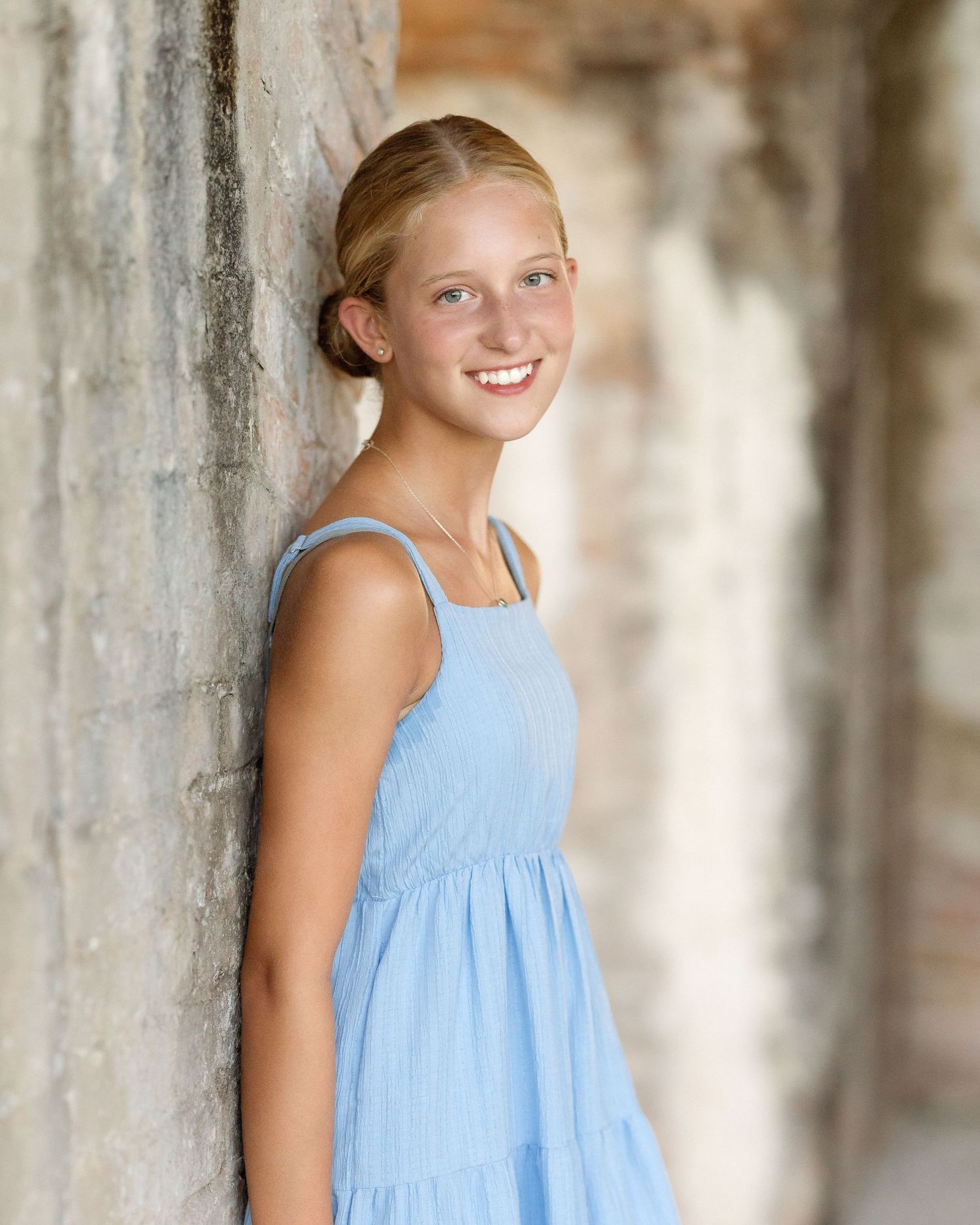 Smiling young girl in a light blue sundress leaning against a stone wall in a bright outdoor setting.