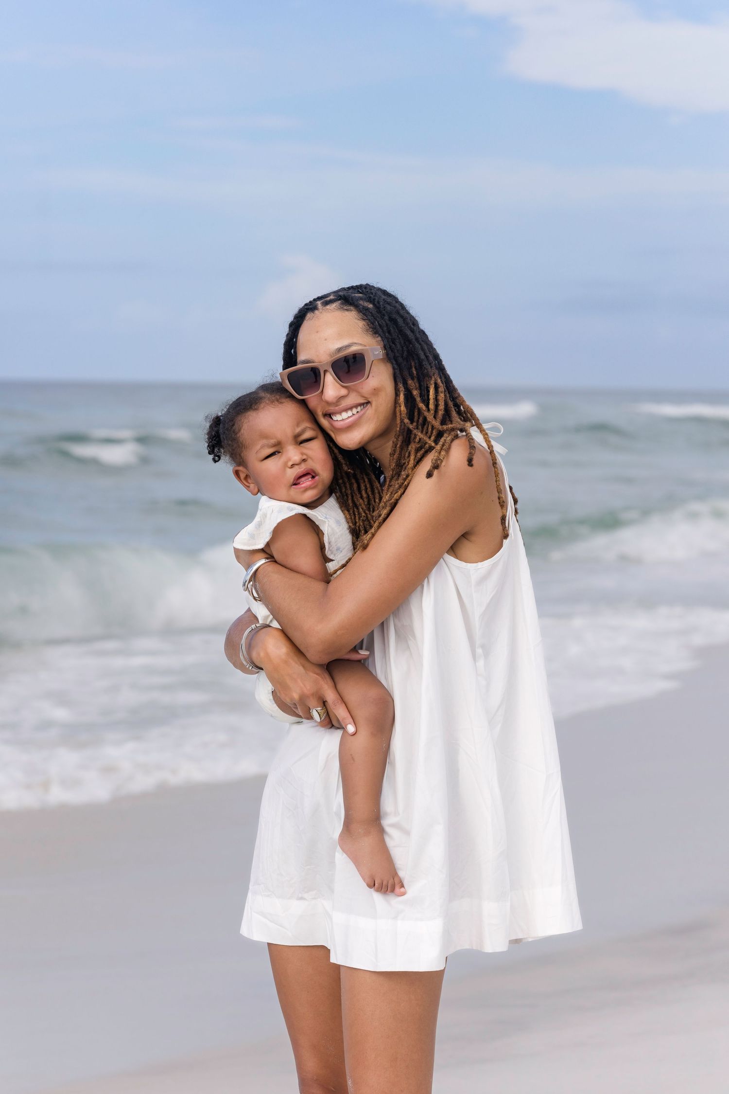 A smiling woman in white dress holds her baby girl on a sunny beach with ocean waves.