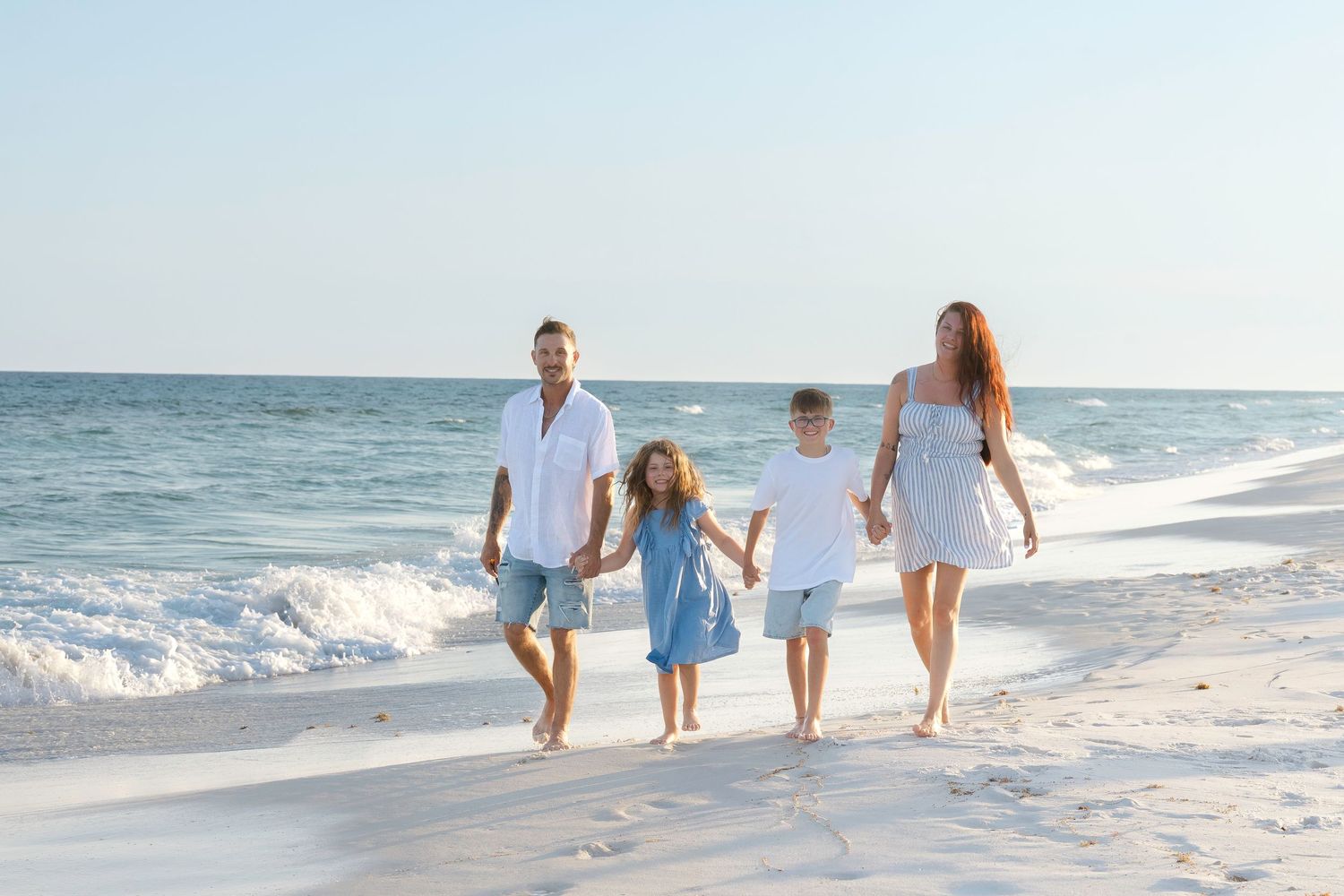 Family of four walking hand-in-hand on a sunny white sand beach with calm turquoise ocean waves in the background.