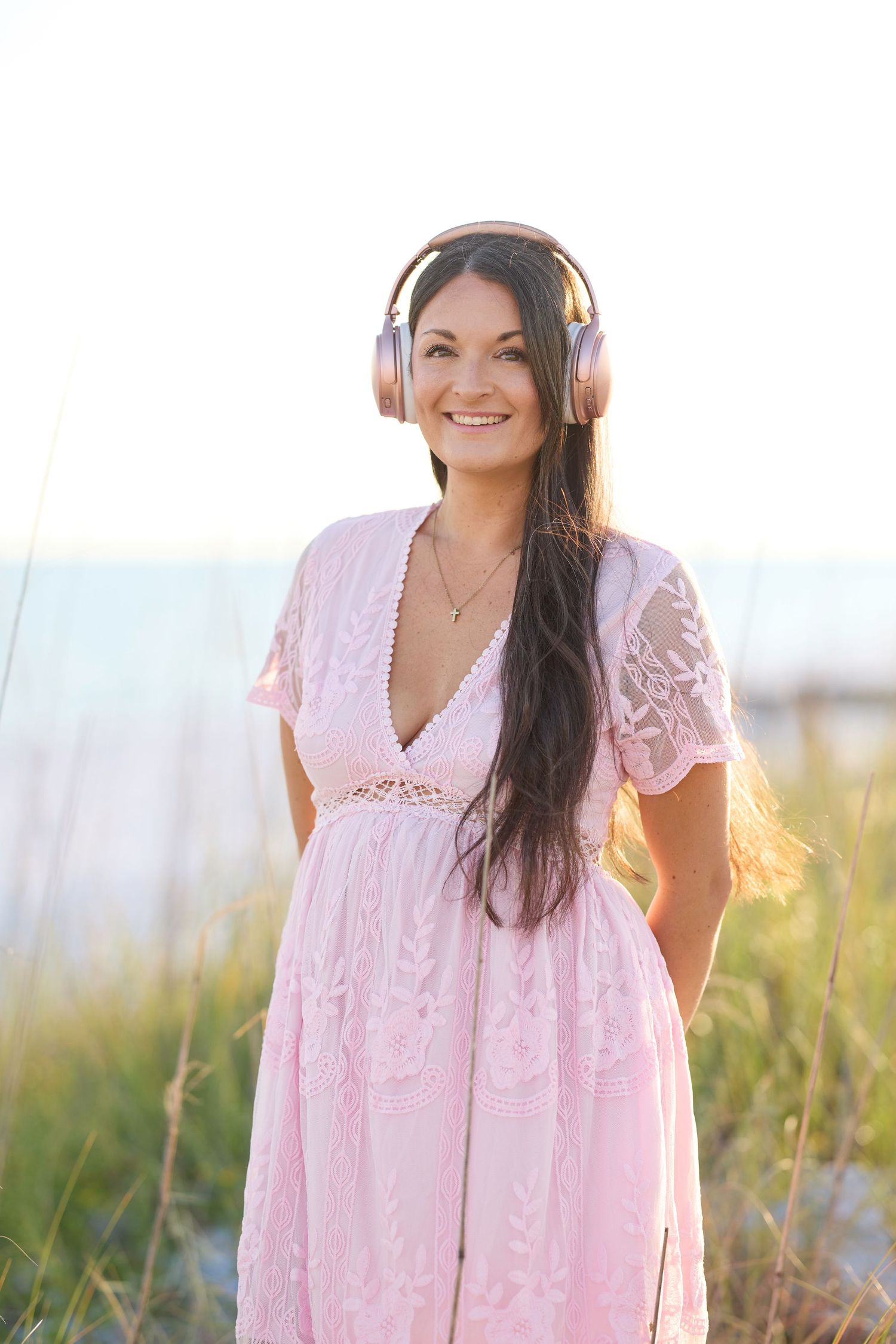 Happy woman in pink lace dress wearing rose gold headphones standing near beach grass at sunset.