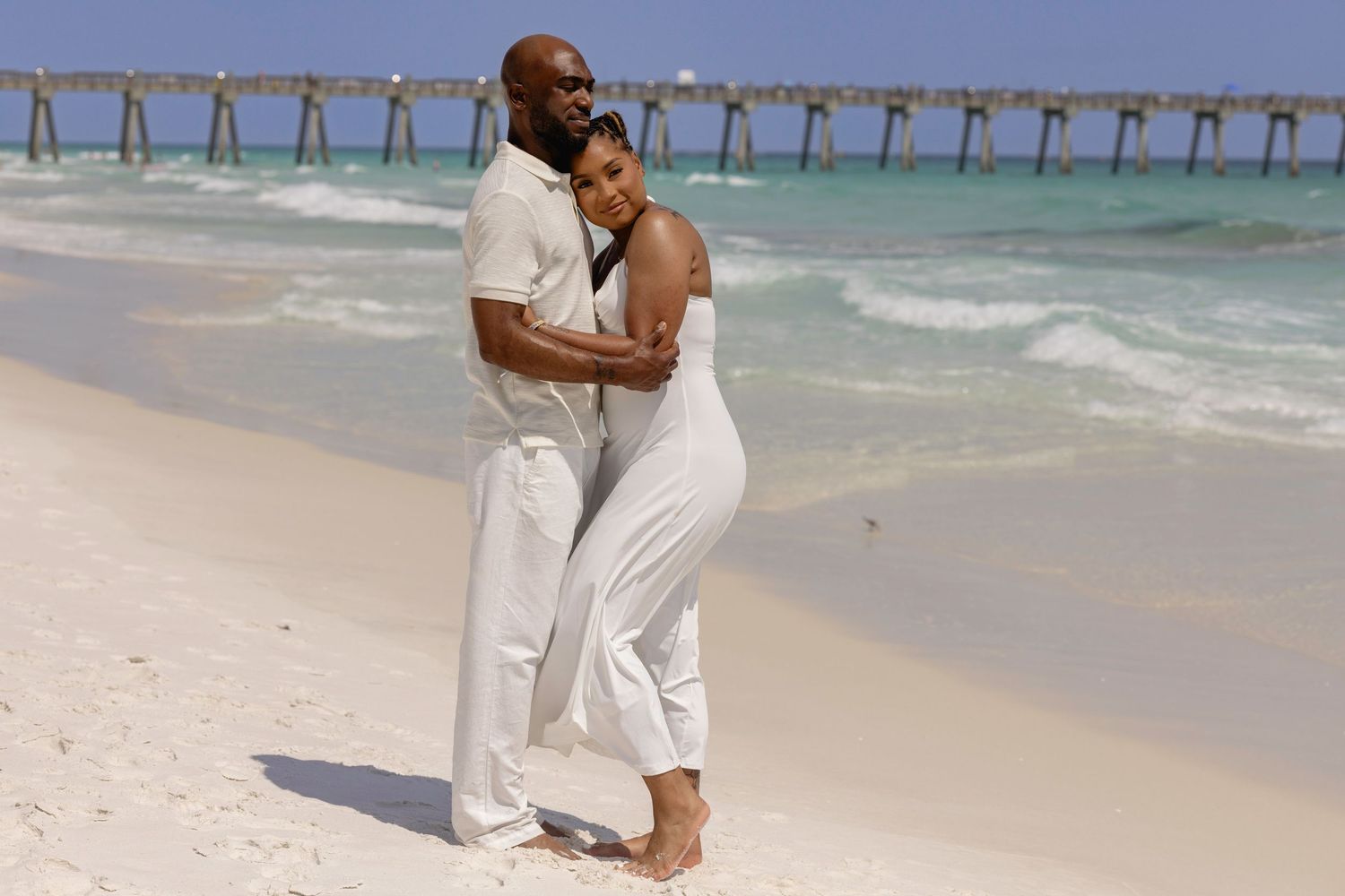 Couple in white outfits embracing on a white sand beach with turquoise water and a pier in the background.