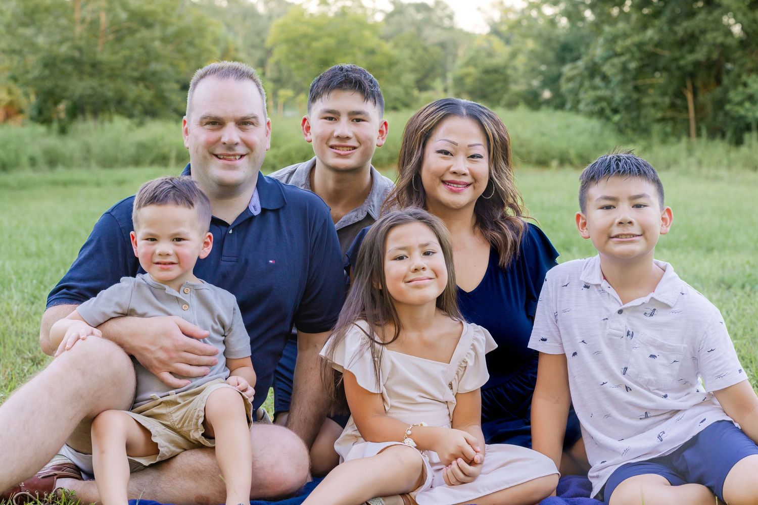 Family of six smiling outdoors on grass, parents with four children posing for a portrait in a lush green park setting.