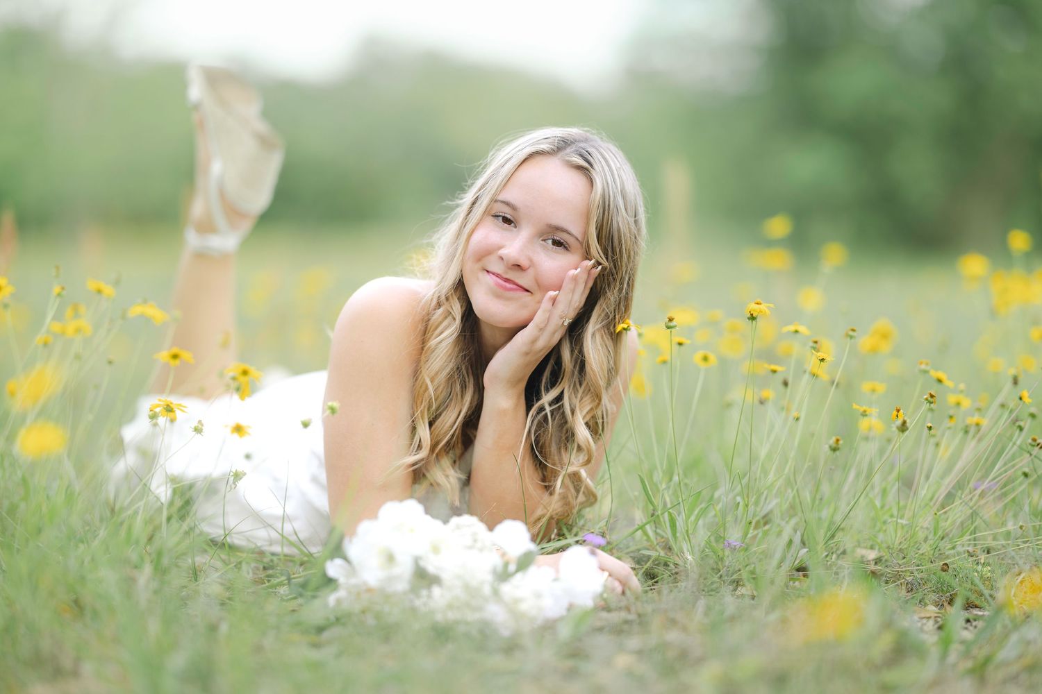 Young blonde woman in white dress lying in a field of yellow wildflowers, smiling softly at the camera.