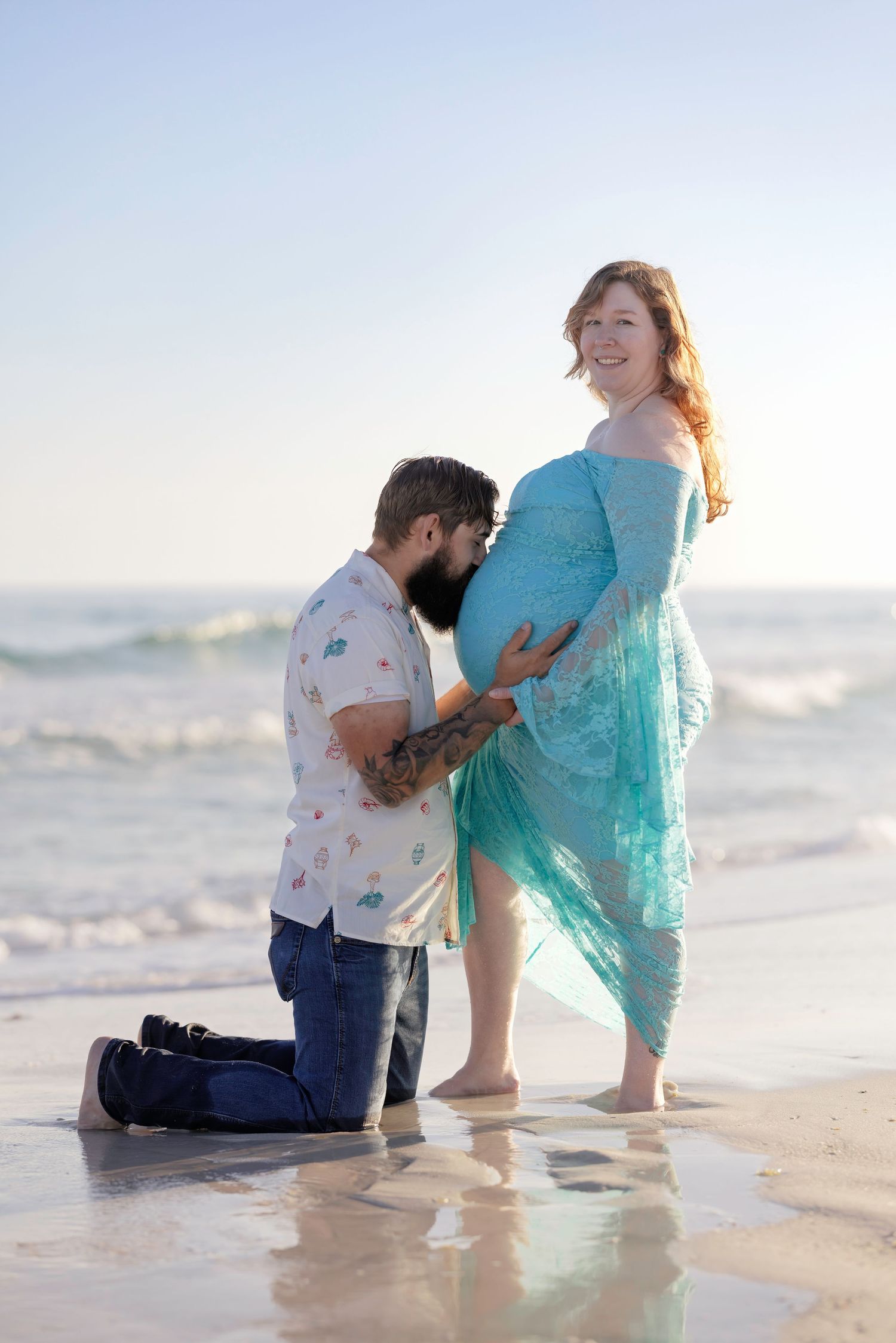 A man kneels on the beach kissing a pregnant woman's belly, as she smiles in a teal lace dress by the ocean.