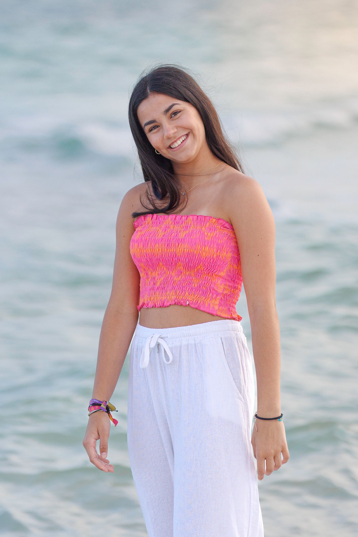 Summer beach portrait featuring coral smocked top and white drawstring pants against ocean backdrop.