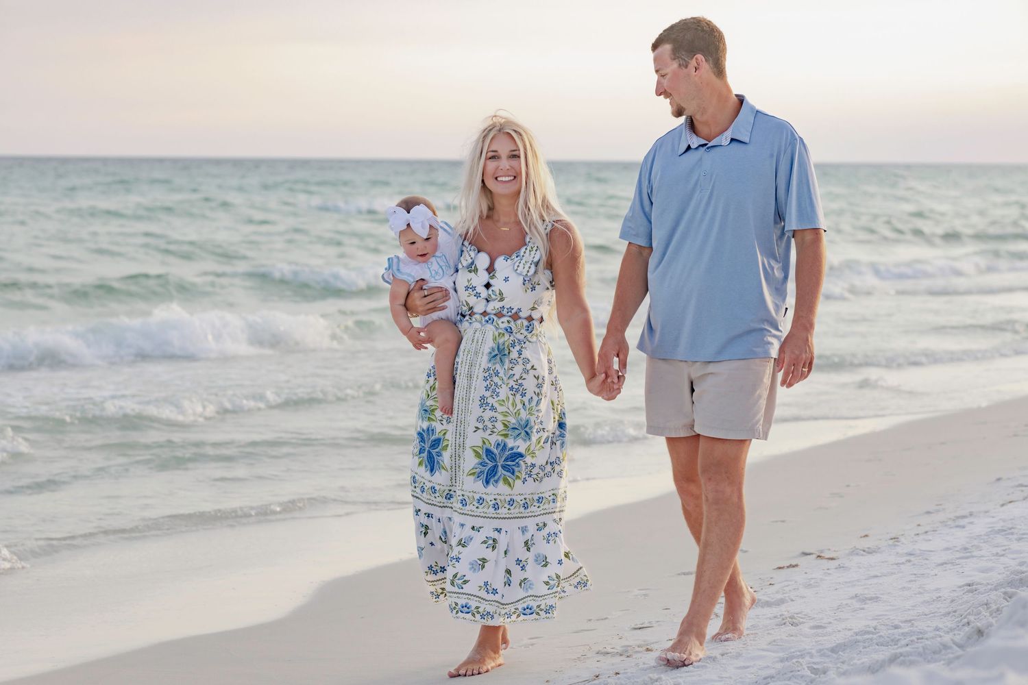 A happy family with a baby walks hand-in-hand on a sandy beach at sunset in casual summer attire.