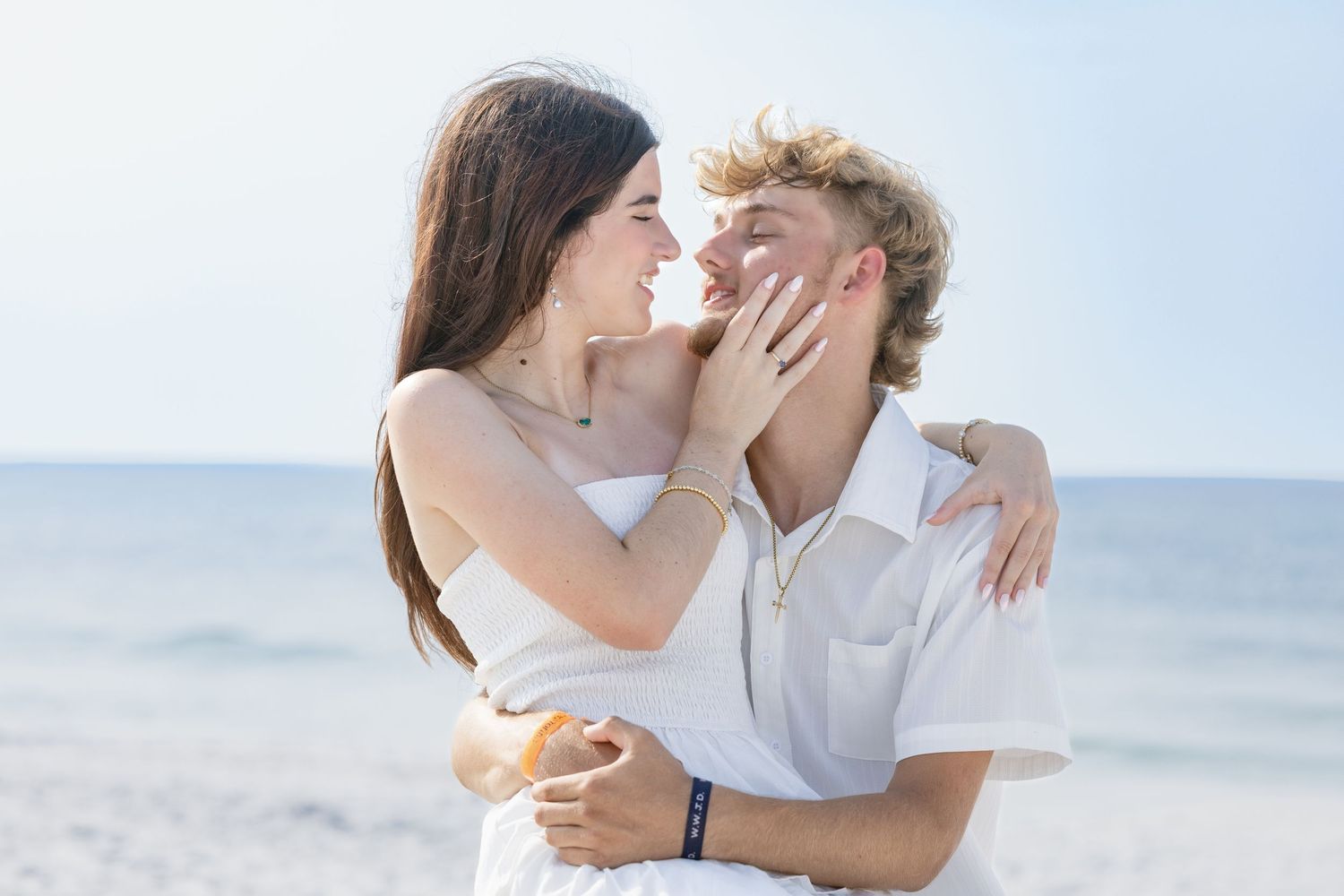 Romantic young couple embracing and almost kissing on a sunny beach, wearing white outfits.