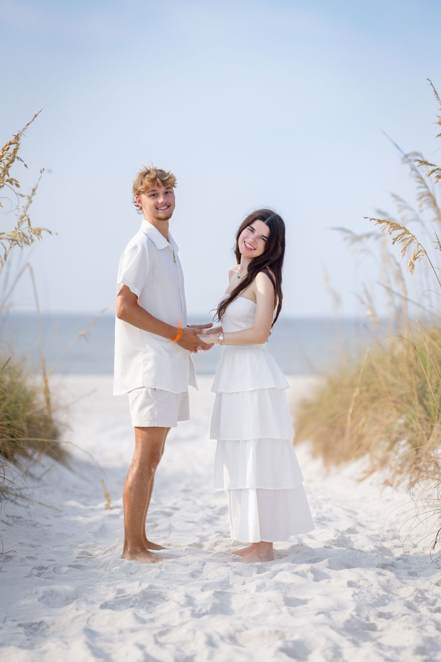 A young couple in matching white outfits holds hands on a sandy beach path surrounded by sea oats.