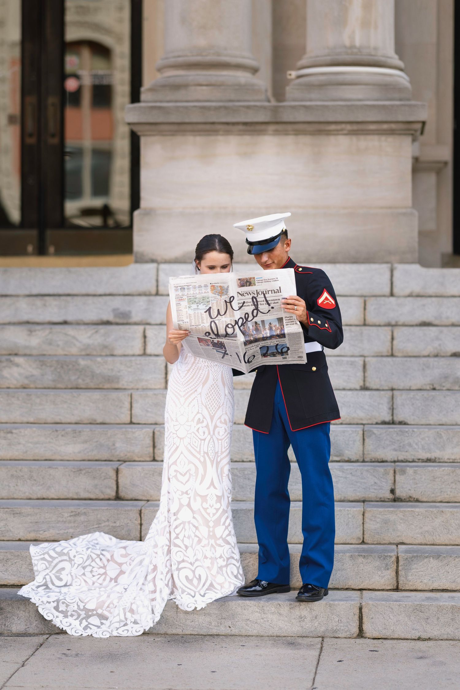 Newlywed Marine groom and bride reading newspaper announcement on steps of historic building.