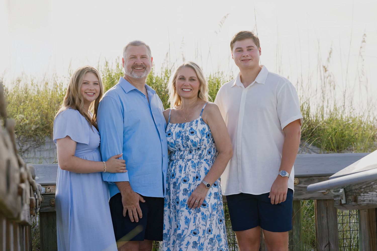 Family of four smiling on a boardwalk near the beach, dressed in blue and white outfits.