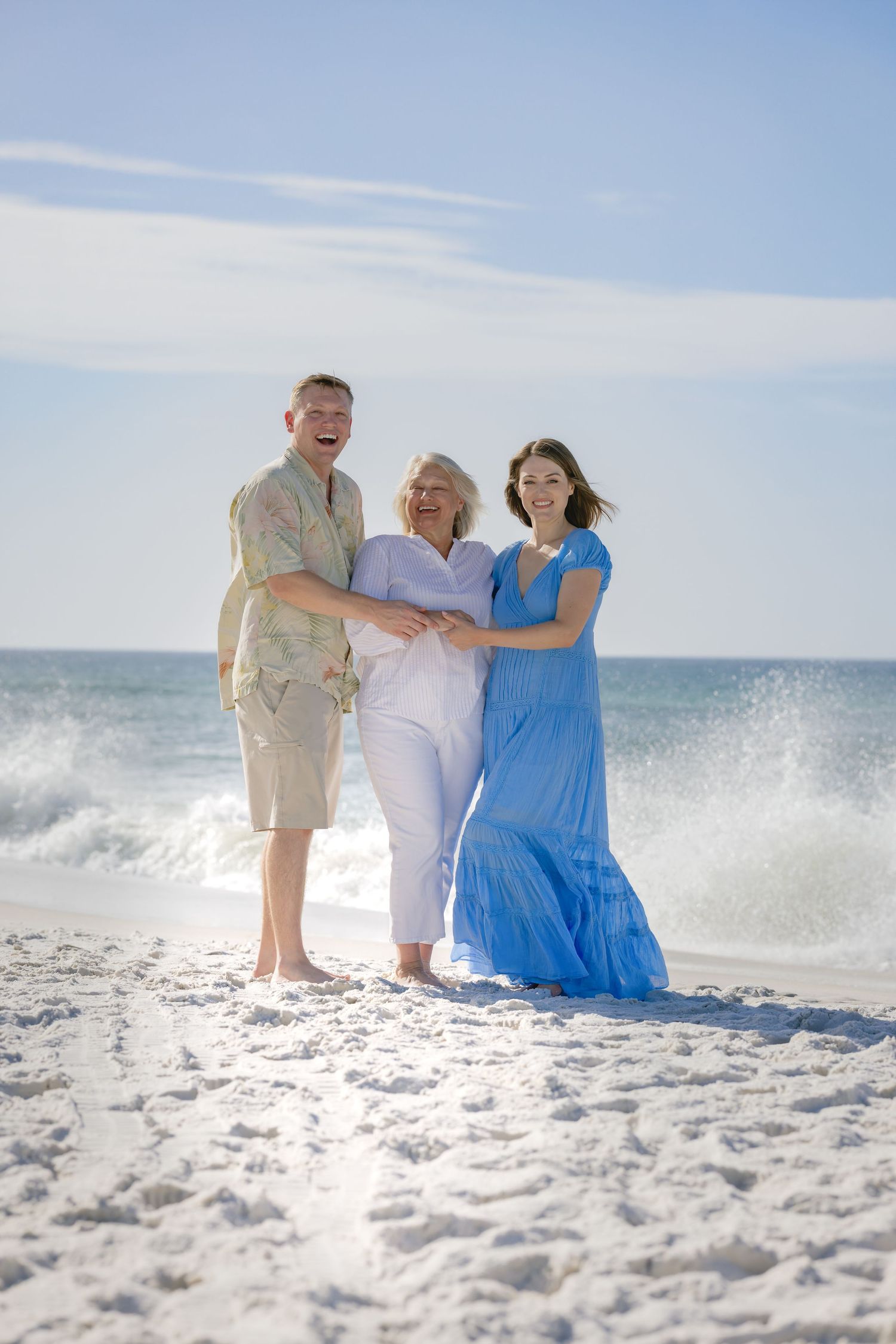 Three happy people pose together on a sunny white sand beach with ocean waves crashing behind them.