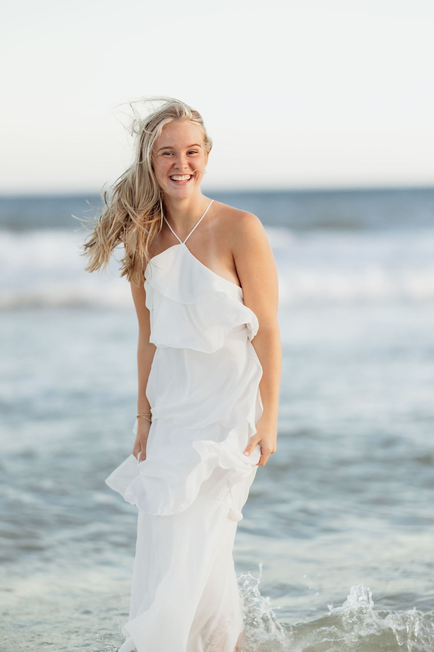 Smiling blonde woman in white dress standing at beach shoreline with ocean waves behind her.