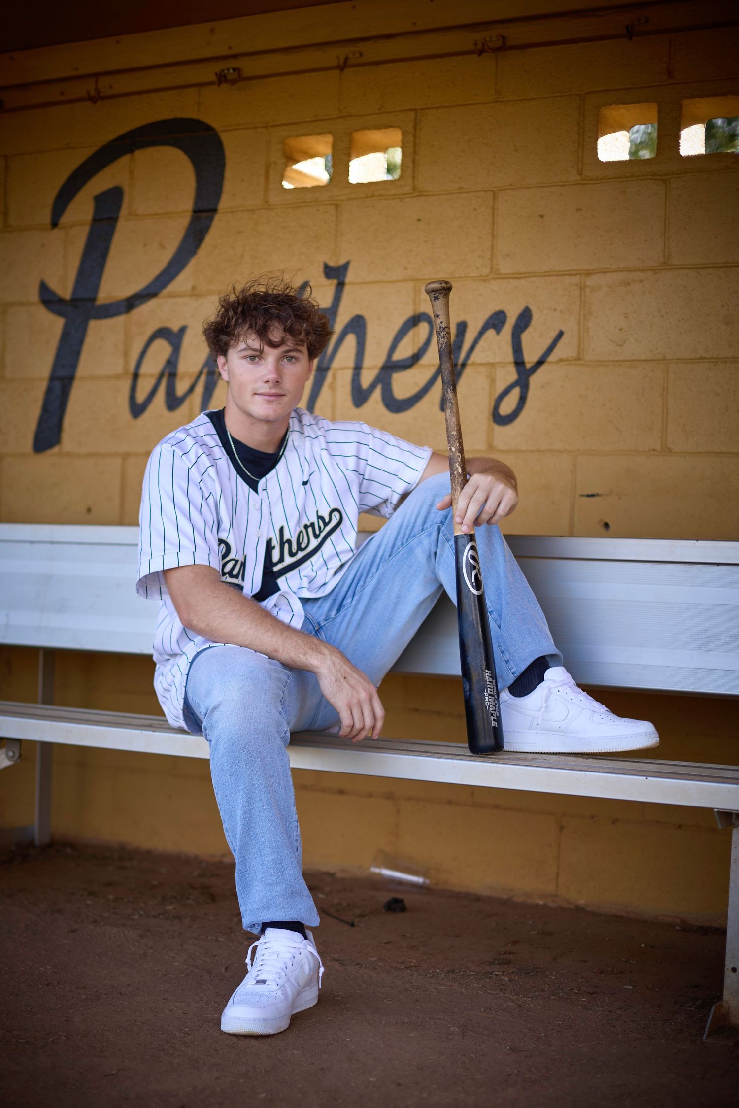 Young baseball player in Panthers jersey sits in dugout holding bat, wearing jeans and white sneakers.