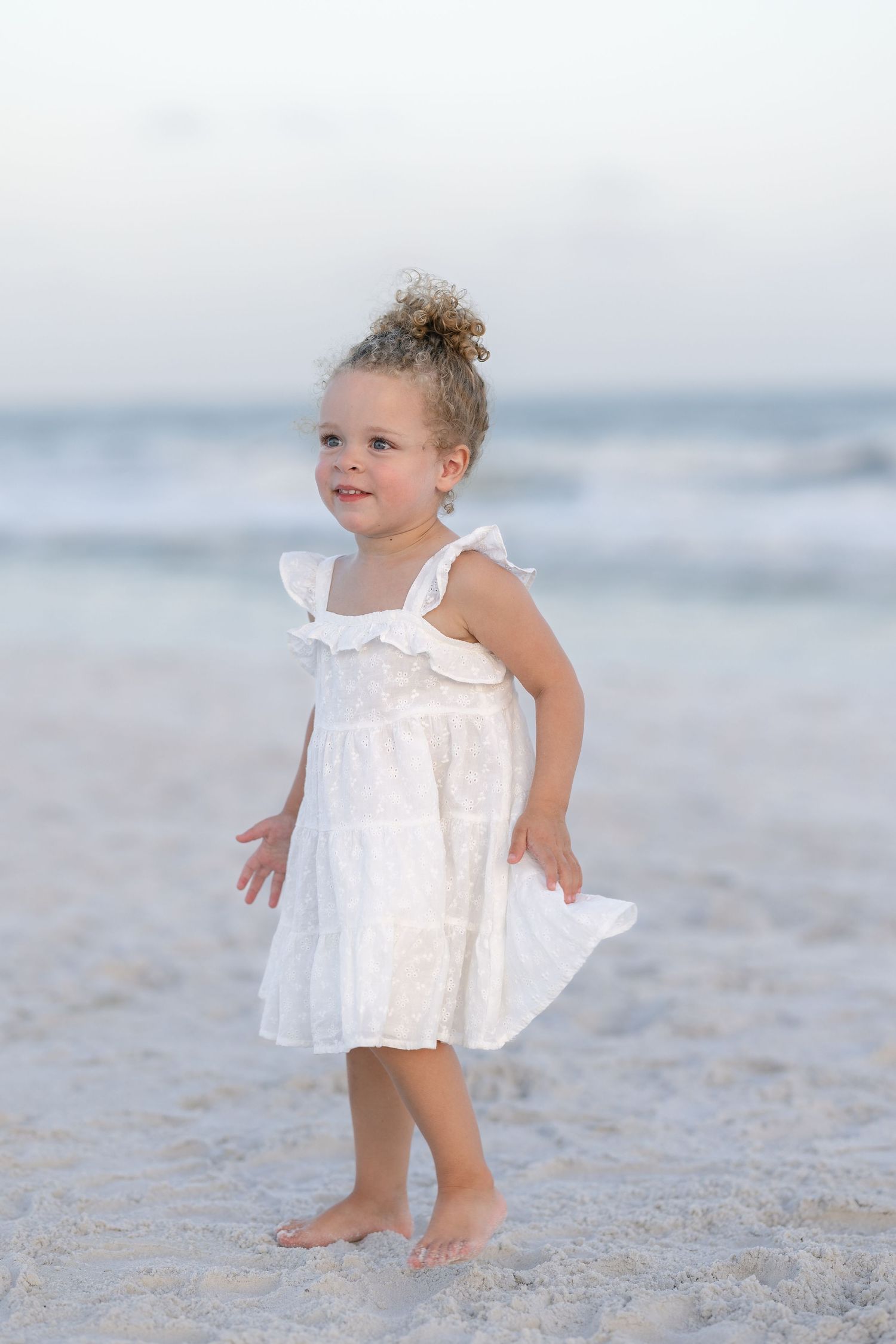 Adorable toddler girl in white dress standing barefoot on sandy beach with ocean waves in background.