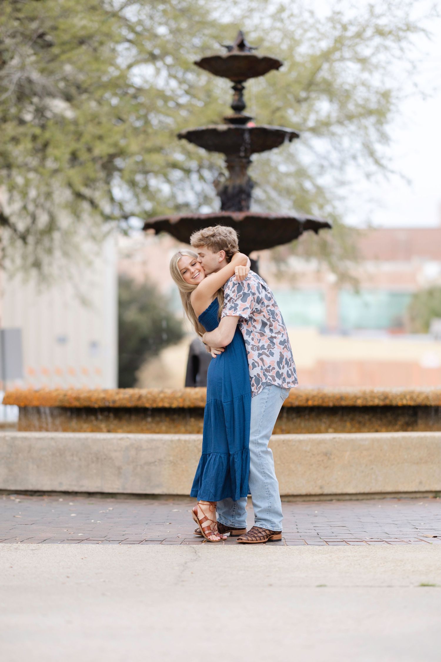 A romantic couple embraces in front of a tiered fountain in an outdoor plaza setting.