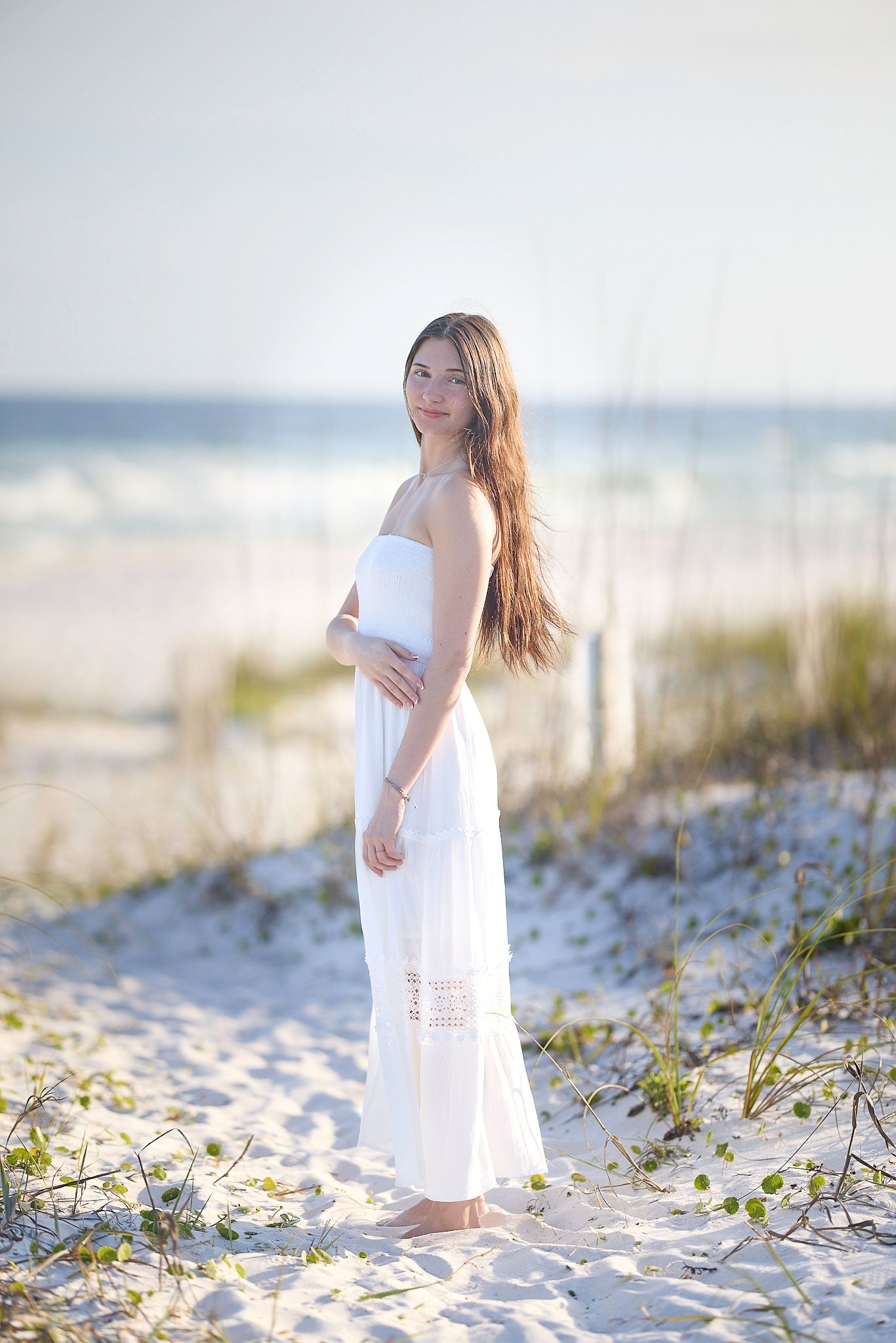 A white sundress flows in the breeze as someone poses on a sandy beach with sea grass and ocean in the background.