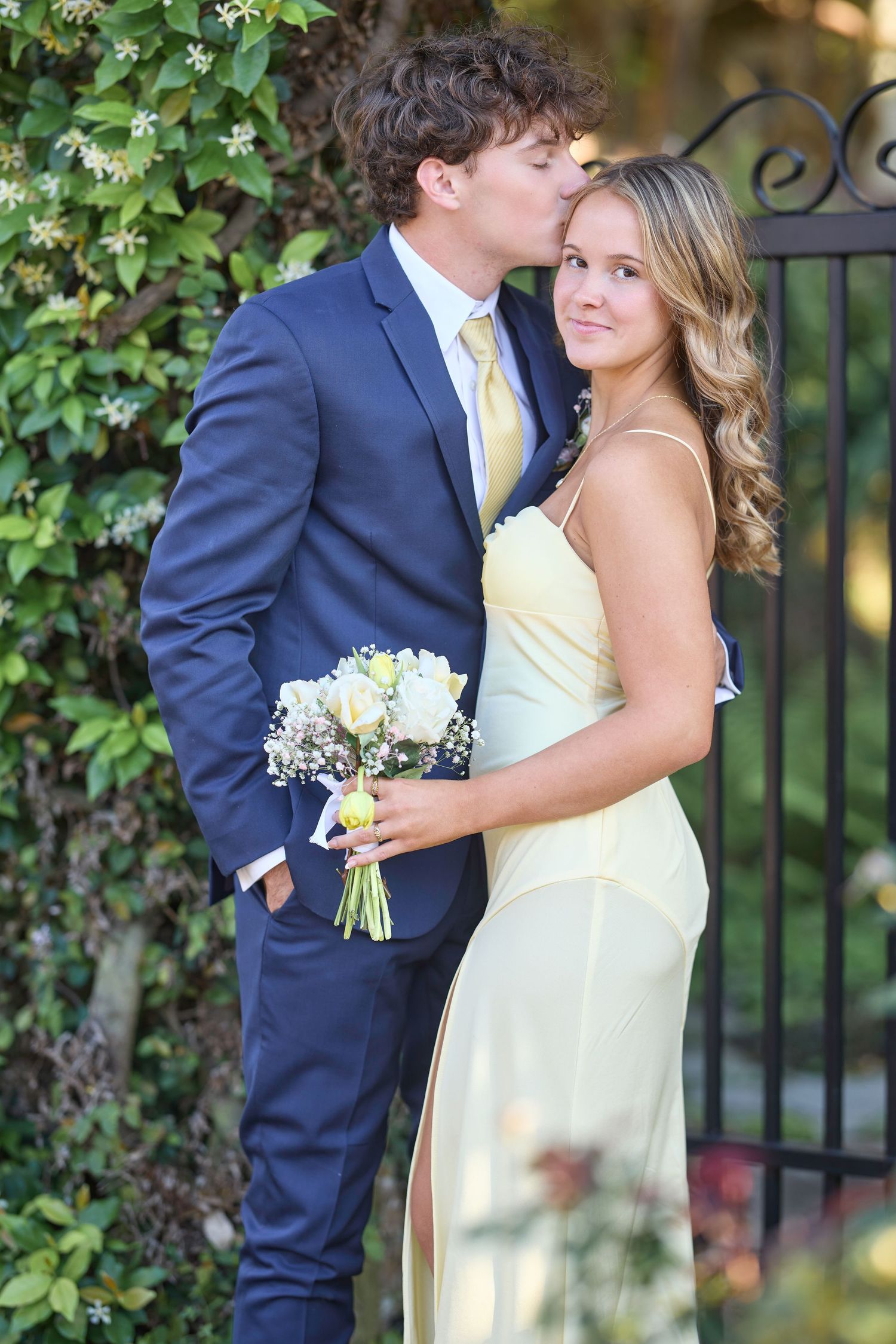 A couple shares an intimate moment in formal attire near an ivy-covered garden fence.