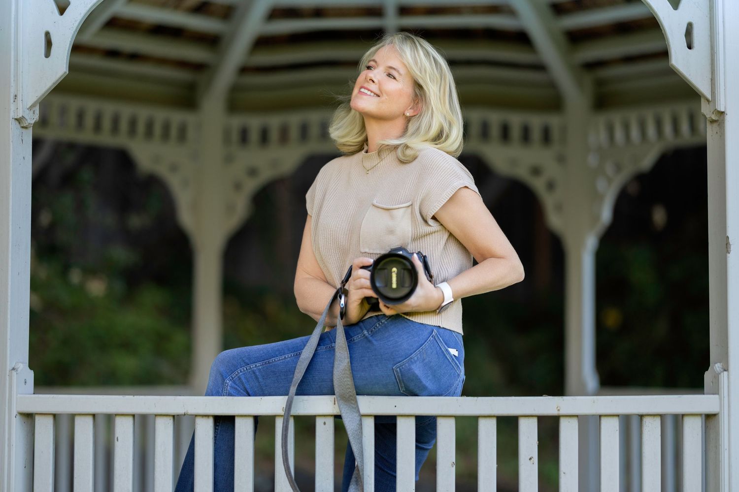 A photographer stands in a white gazebo holding a professional camera while wearing jeans and a beige top.
