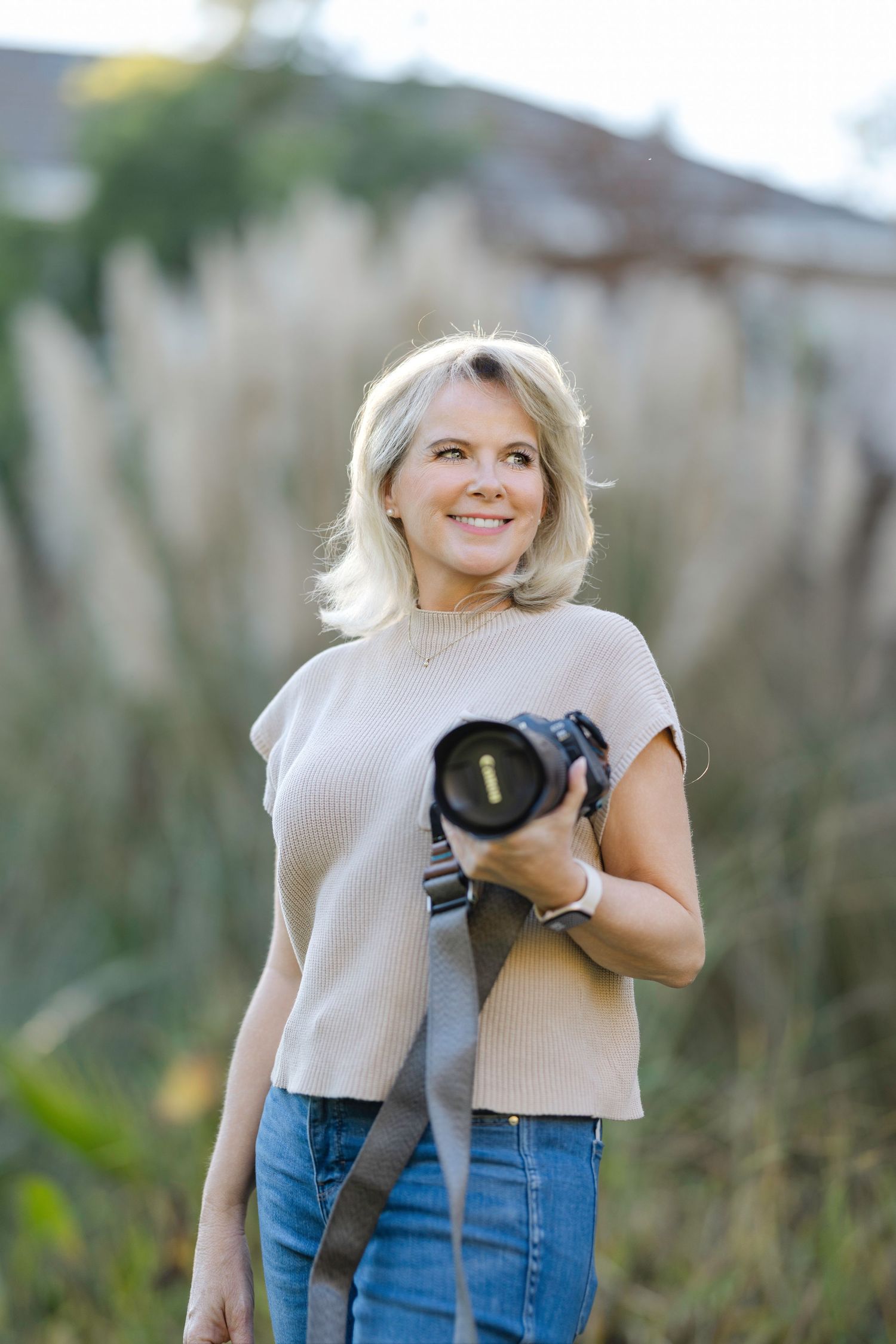Professional photographer in beige sweater and jeans holding camera equipment outdoors.