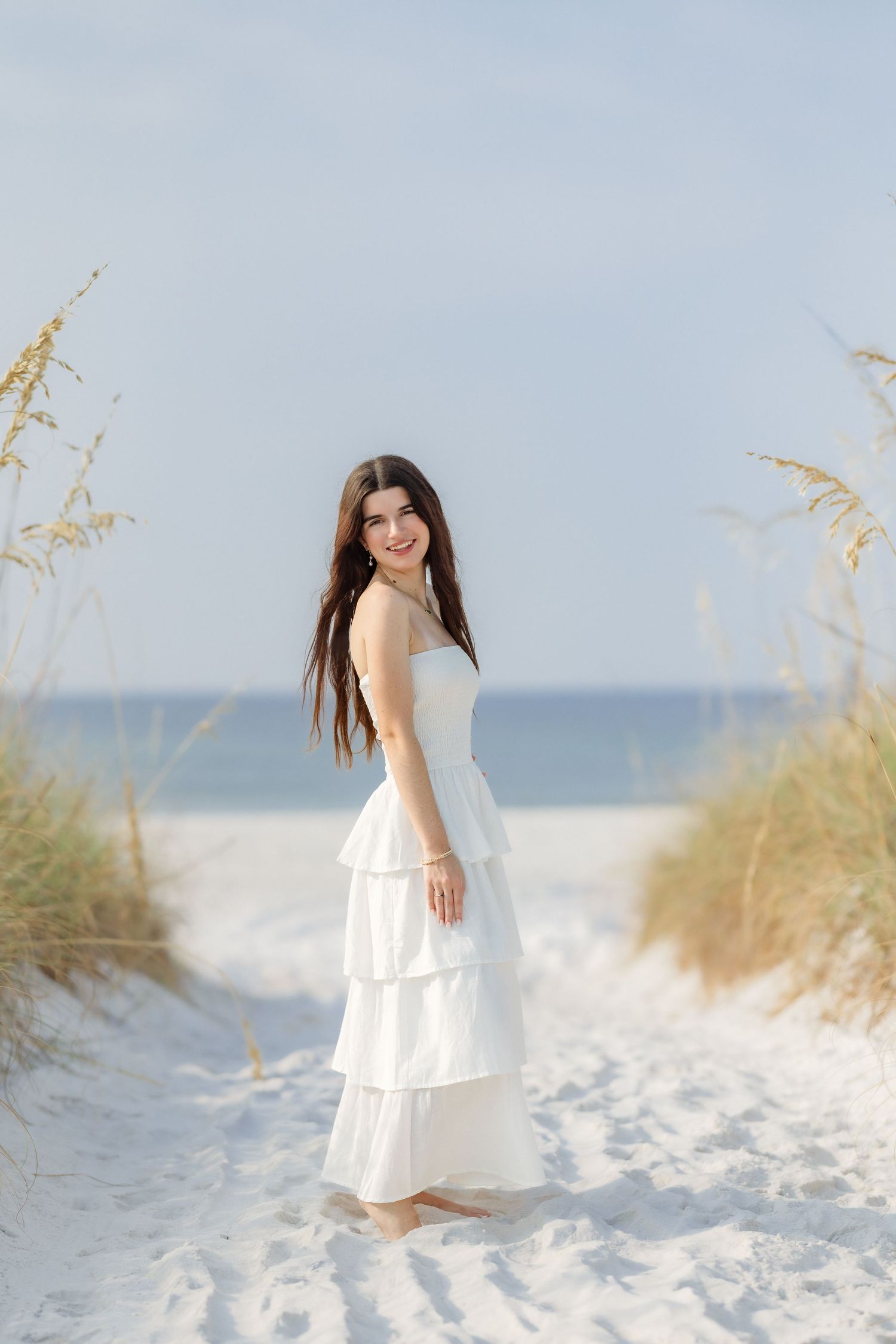 Young woman in white tiered dress standing on sandy beach path with sea oats, smiling over shoulder.