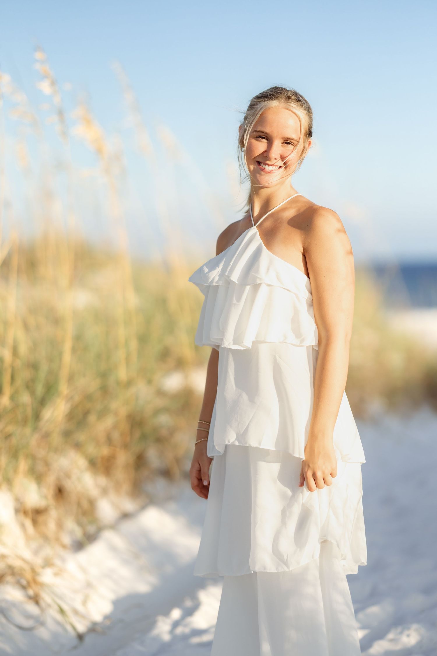Young blonde woman in white tiered ruffle halter dress smiling on white sand beach dunes.