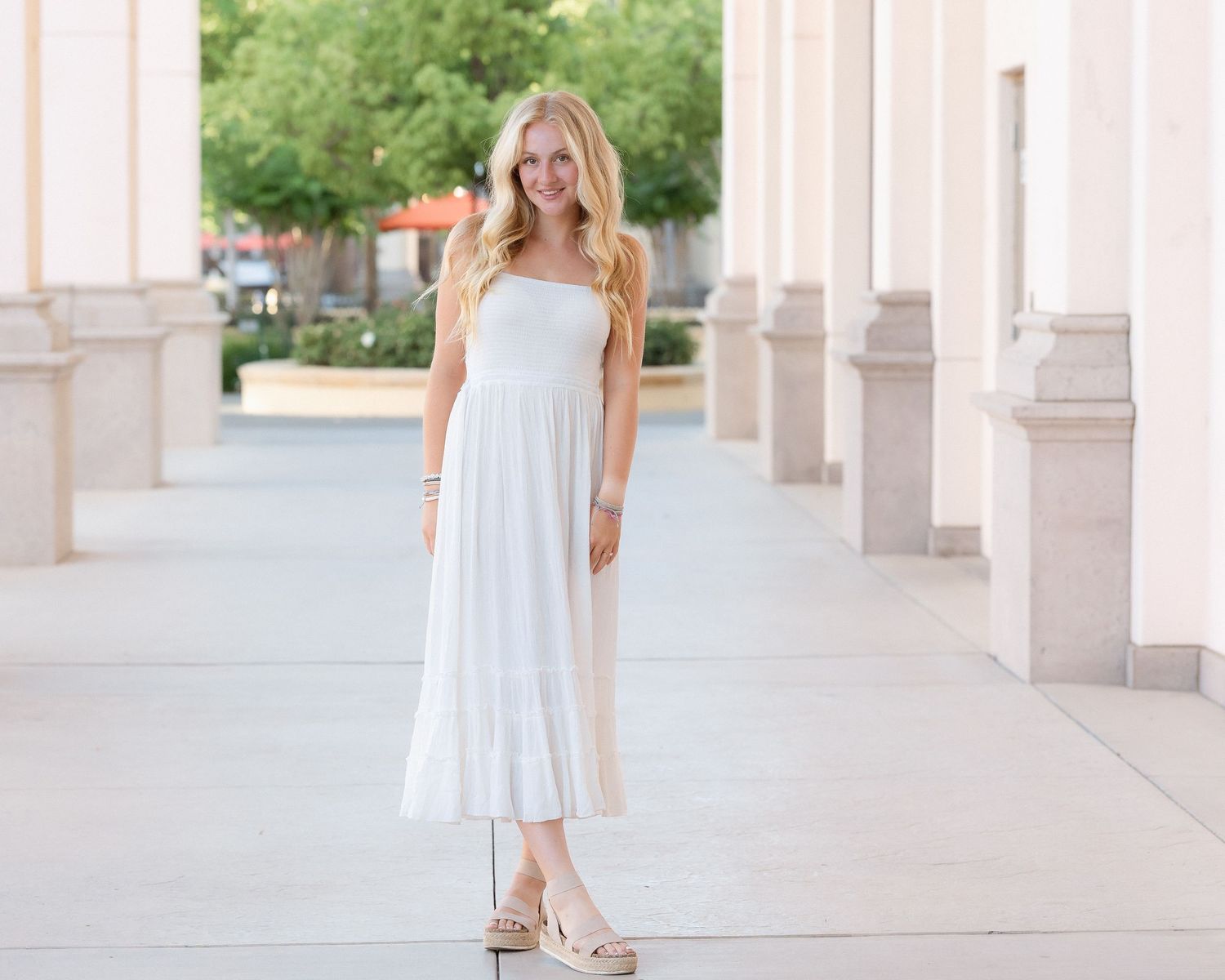 Blonde teenage girl in a white maxi dress poses gracefully between columns outdoors.