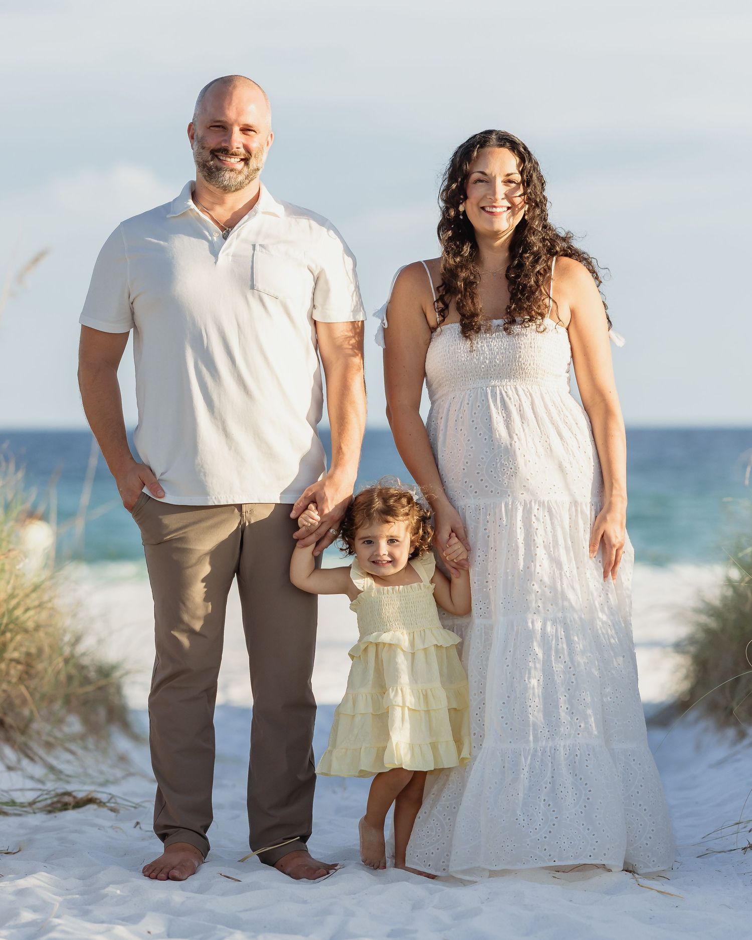 Family of three posing on a sandy beach, parents holding toddler girl's hands, wearing white and neutral tones.