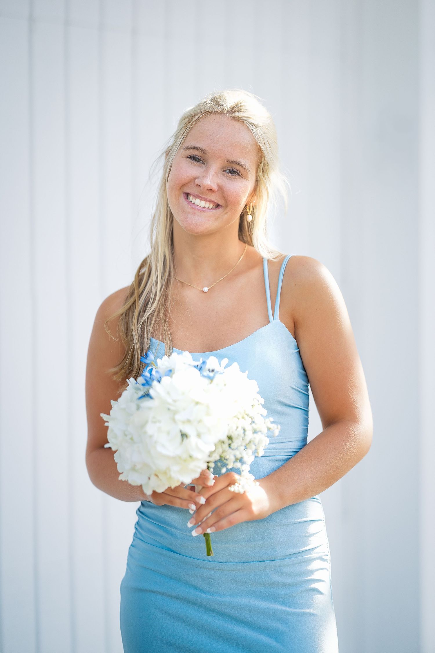 Bridesmaid in light blue dress holding white floral bouquet poses against light background.