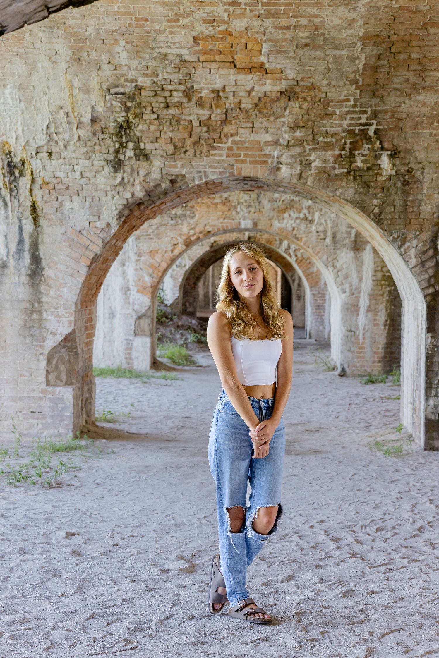 A casual summer outfit featuring ripped jeans and a white crop top under historic brick archways at a fort.