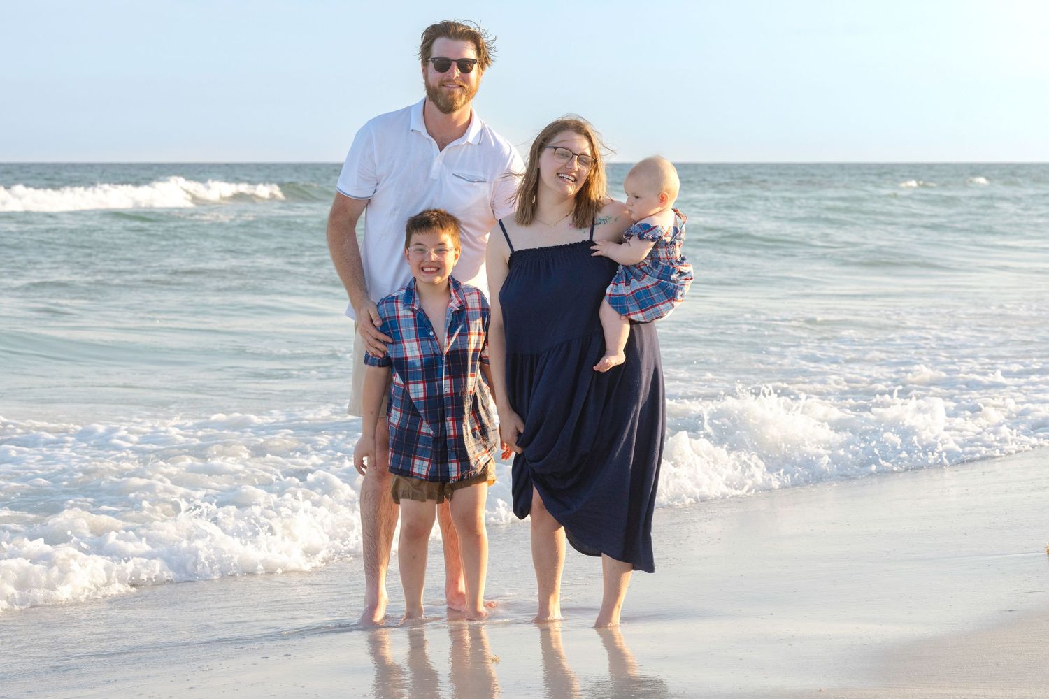 Happy family of four standing at ocean's edge on a sunny beach, mother holding baby, son beside father.