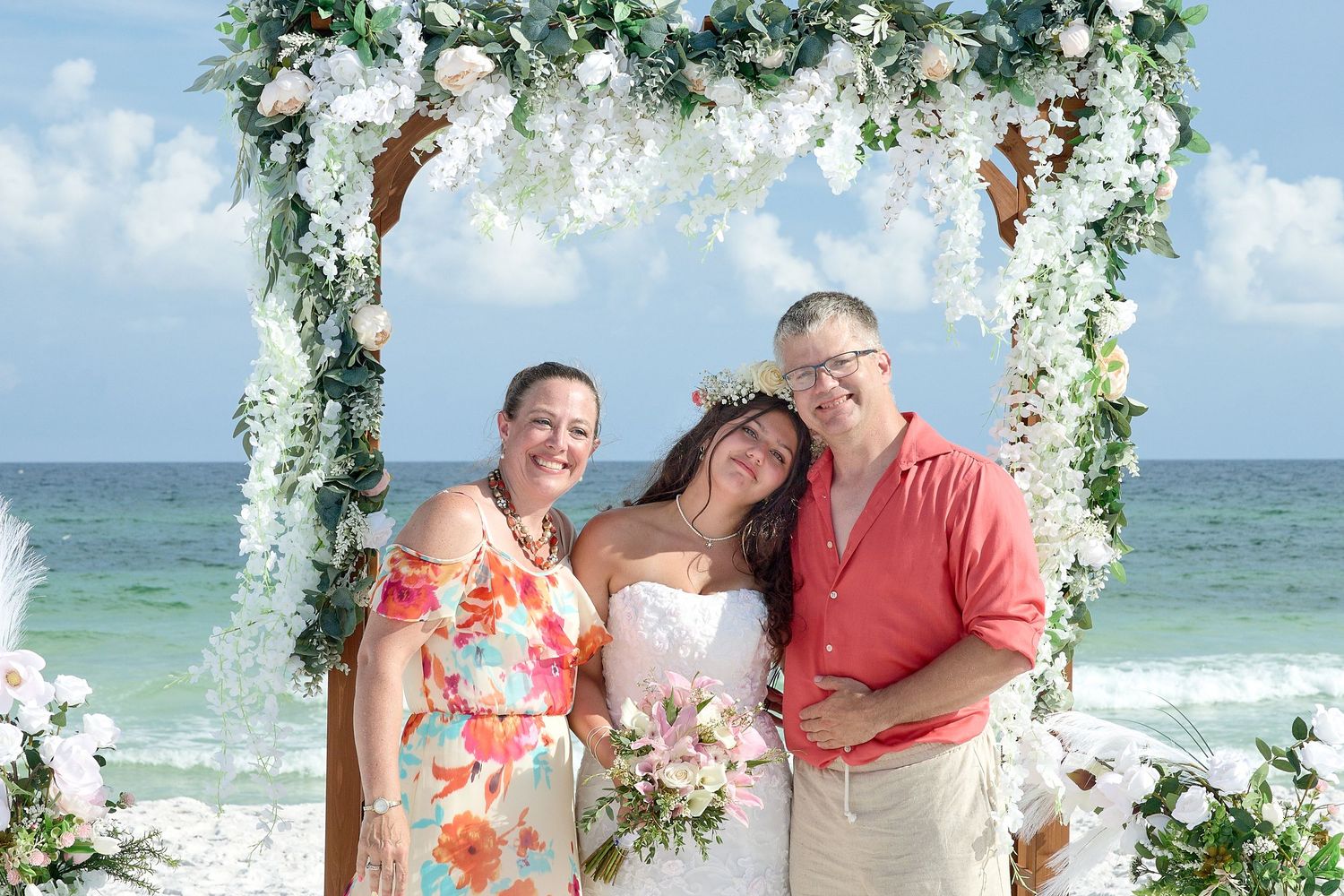 Bride in white gown with bouquet poses under floral arch on beach with two guests at outdoor wedding ceremony.