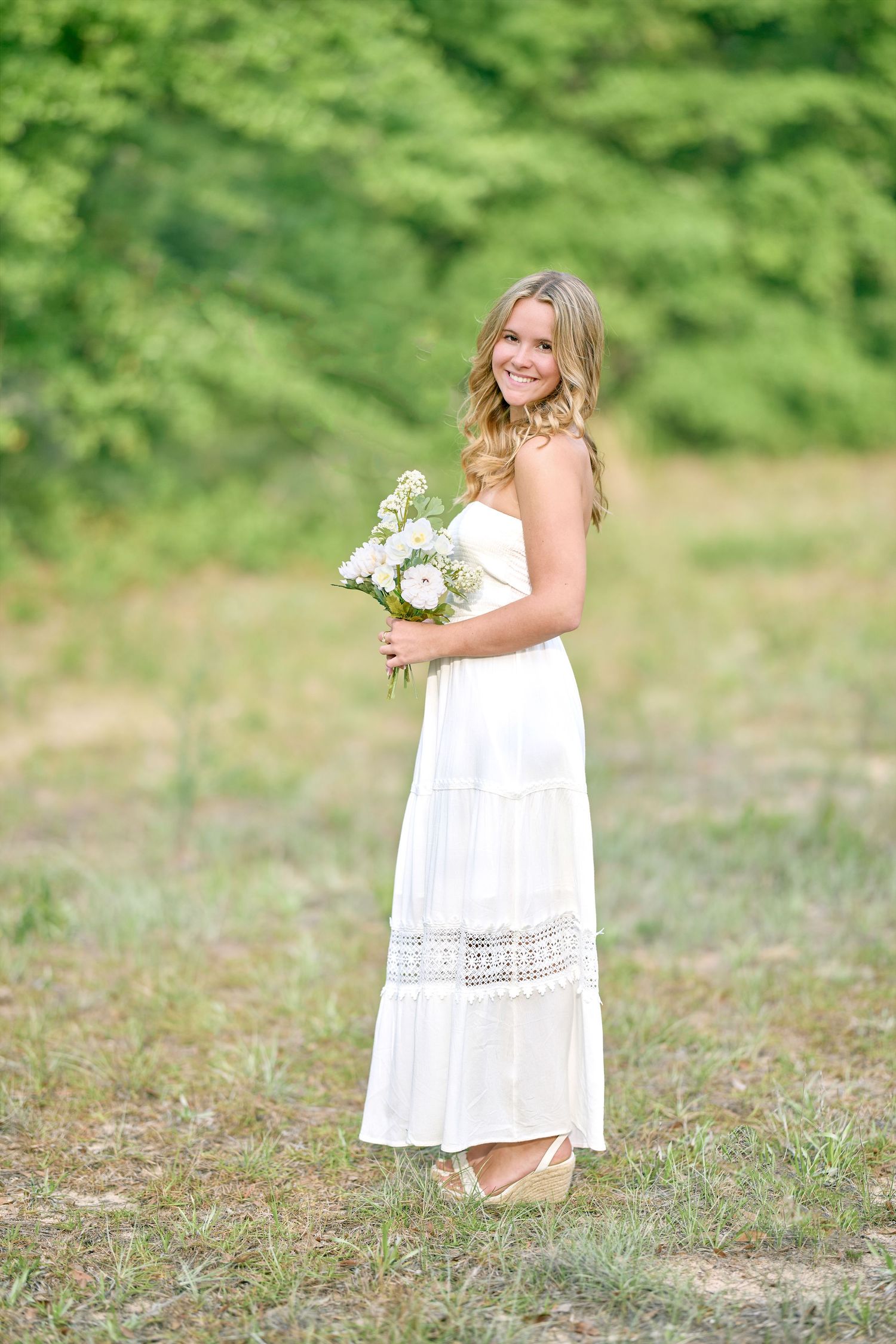 Full length portrait in flowing white maxi dress holding floral bouquet.
